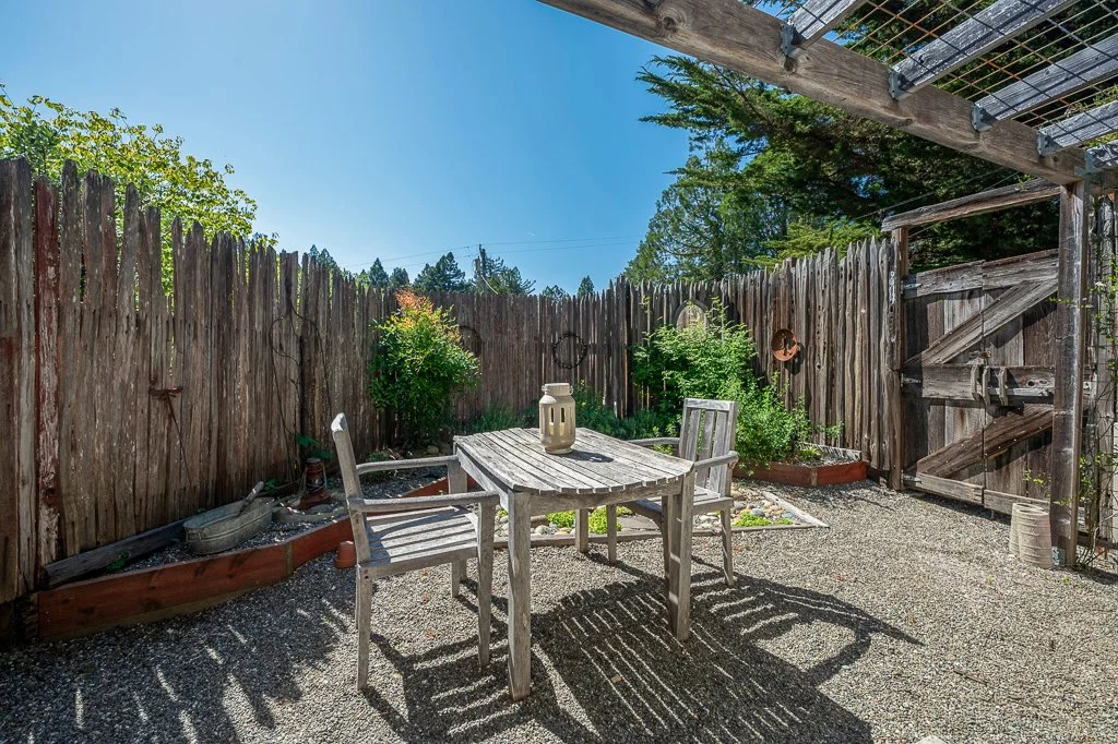 A small outdoor patio area enclosed by a weathered wooden fence, with a gravel ground, a wooden table with three chairs, and some greenery in raised garden beds and planters, under a clear blue sky.