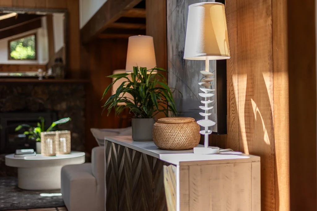 Interior of a cozy living room with a wooden wall, two table lamps, a houseplant, a wicker basket, and a white cabinet, illuminated by natural sunlight.