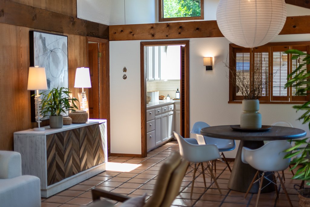 Living room with wooden accents, a round dining table with white chairs, a sideboard with lamps and plants, and a view into the kitchen.