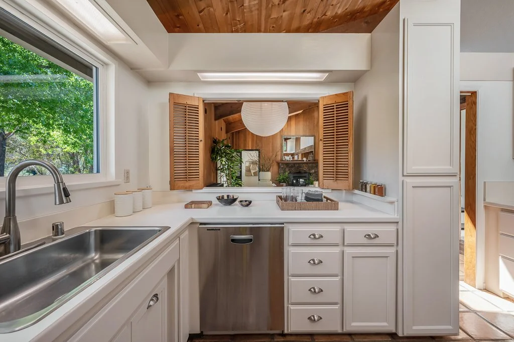 Kitchen with white cabinets, stainless steel dishwasher, double sink, window showing green trees outside, wooden ceiling, pass-through window with shutters, and a view into a dining area with wood-paneled wall, plants, and a paper lantern light.