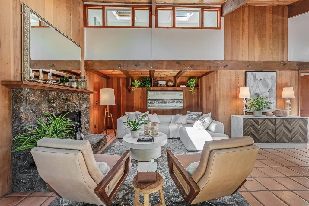 Living room with wood-paneled walls, a white sectional sofa, two beige armchairs, a stone fireplace, a round coffee table, and decorative plants.
