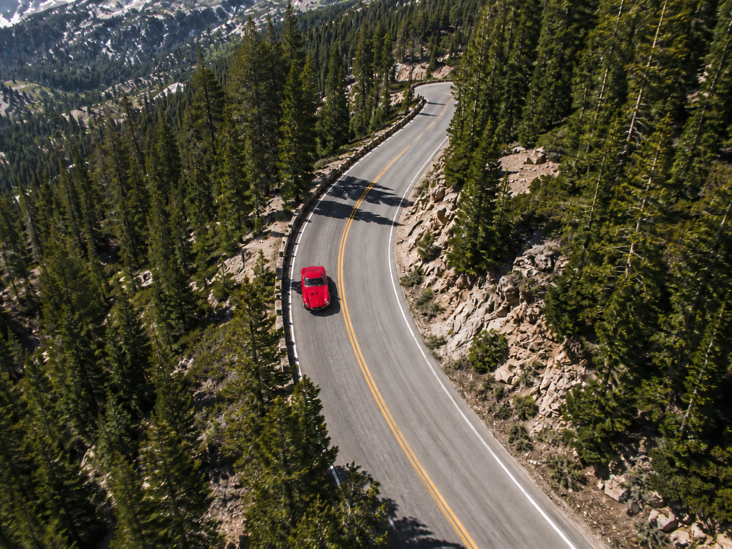 A red car driving on a winding mountain road surrounded by dense forest of pine trees.