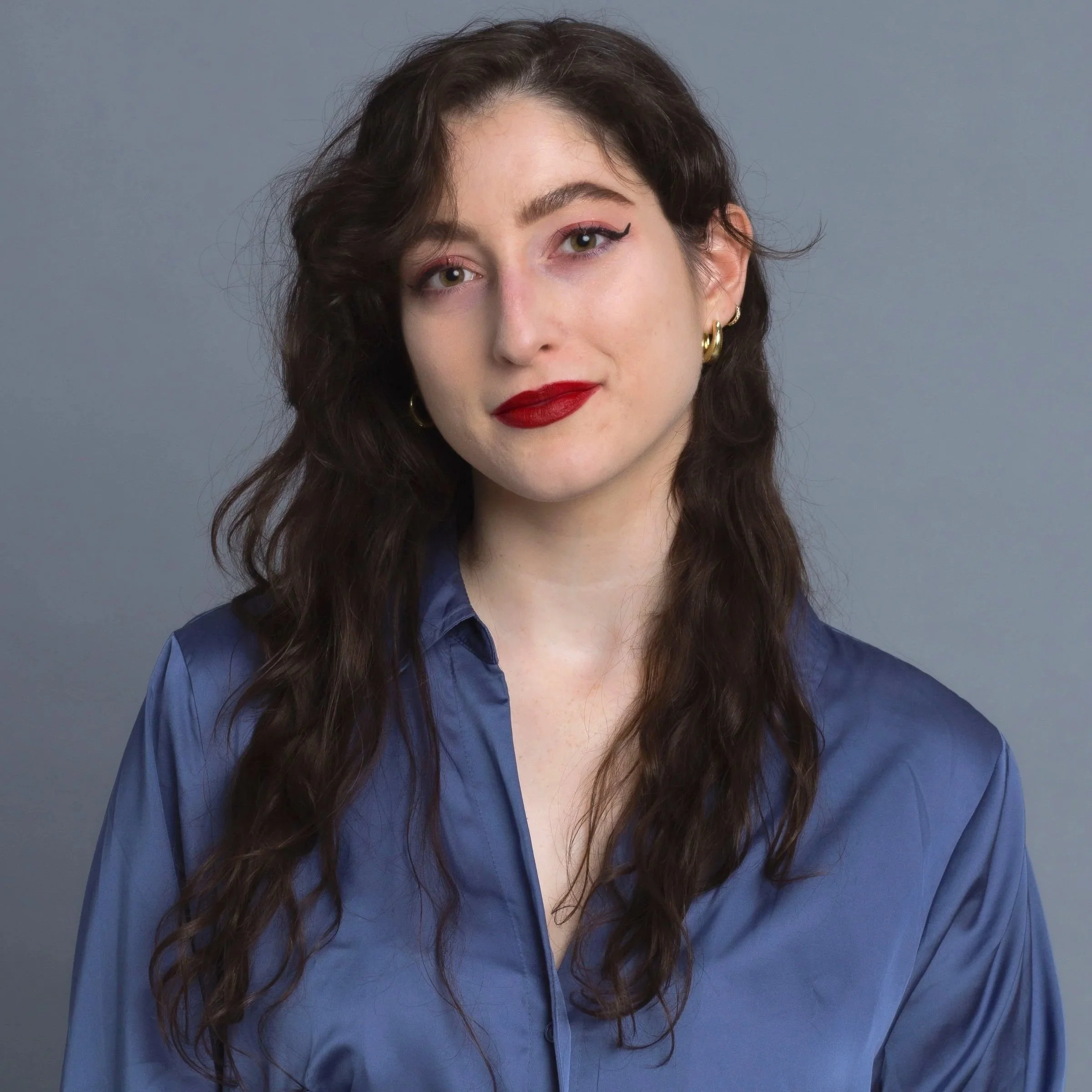 Close-up of a woman with dark wavy hair, wearing gold hoop earrings, red lipstick, eyeliner, and a blue shirt, against a gray background.