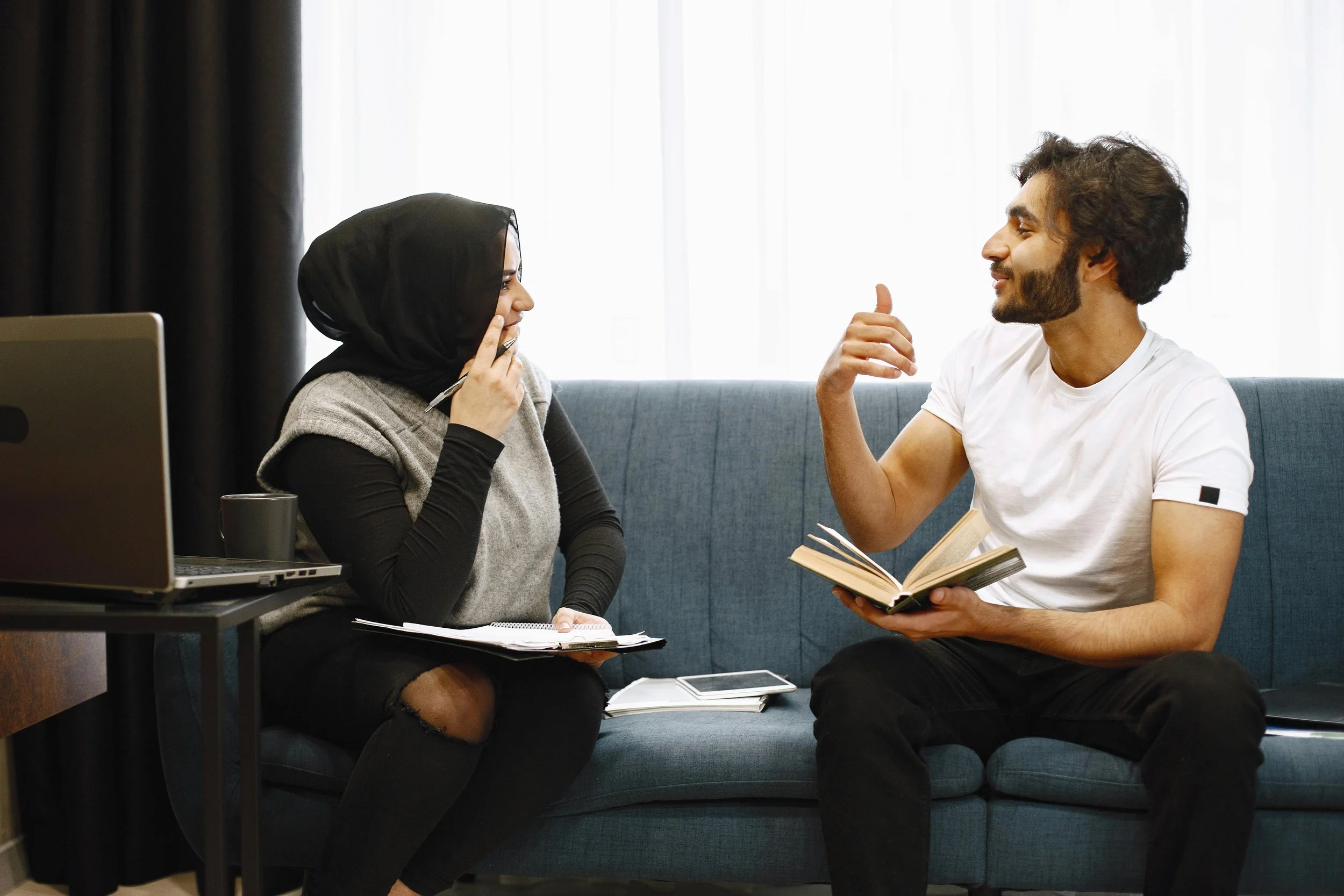 Two people sitting on a blue couch engaged in a conversation, with a woman wearing a black hijab and a man with curly hair and a beard holding a book while gesturing.