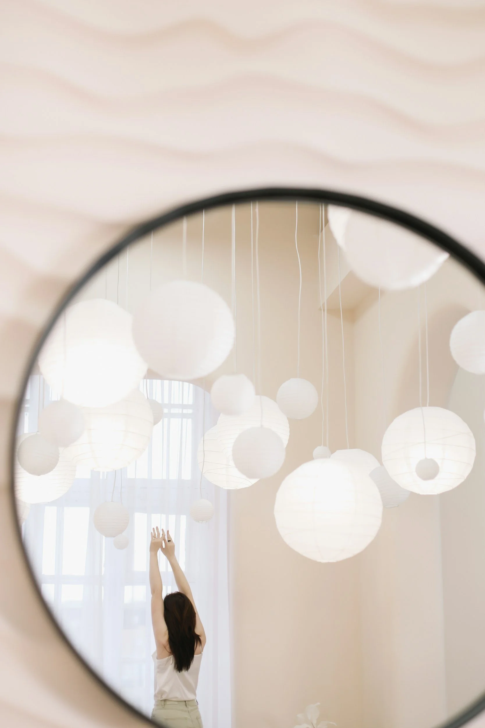 A woman with long dark hair is reaching up with both hands in a room with white paper lanterns hanging from the ceiling.