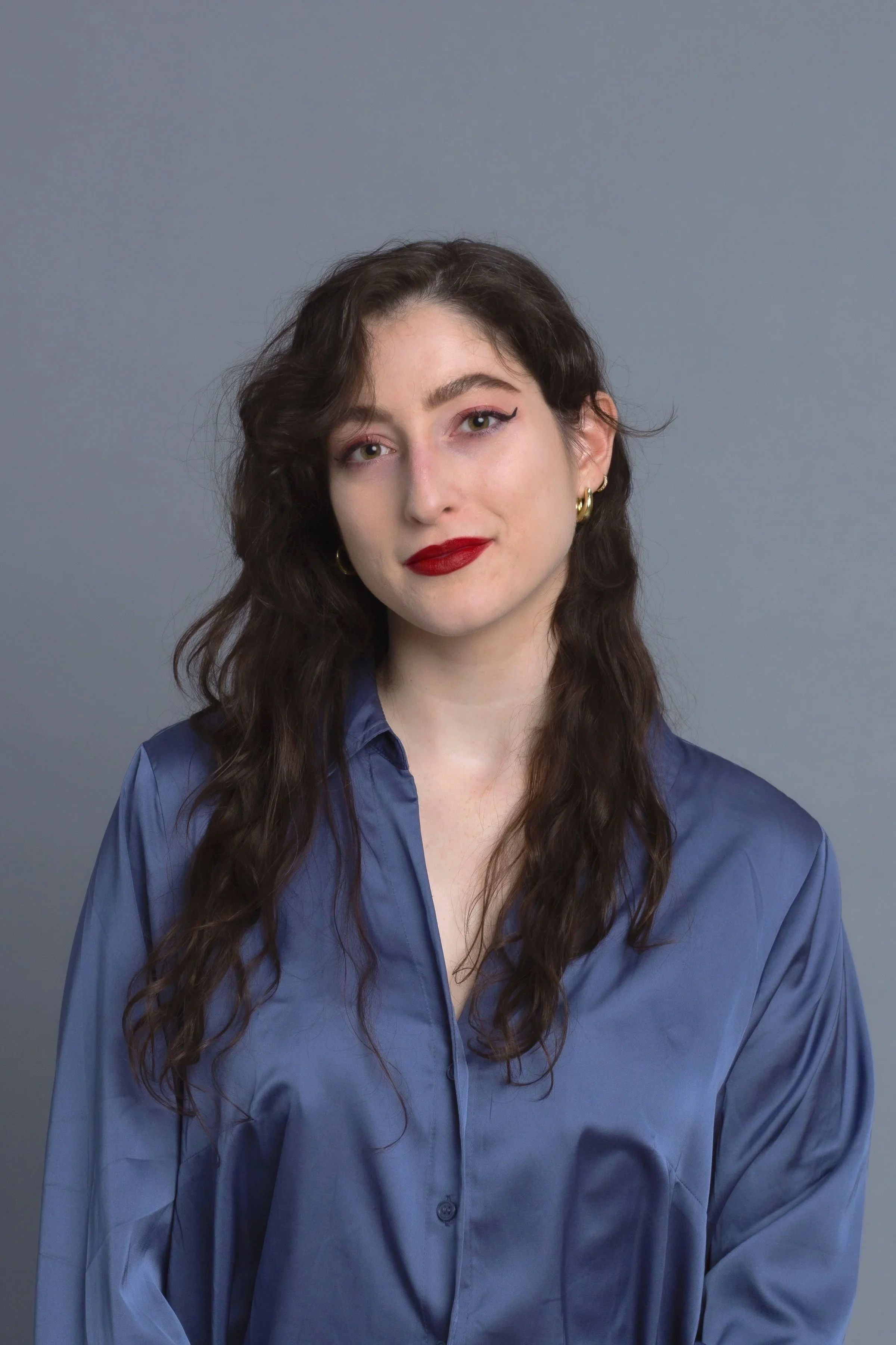A woman with dark wavy hair wearing a blue button-up shirt against a plain gray background.