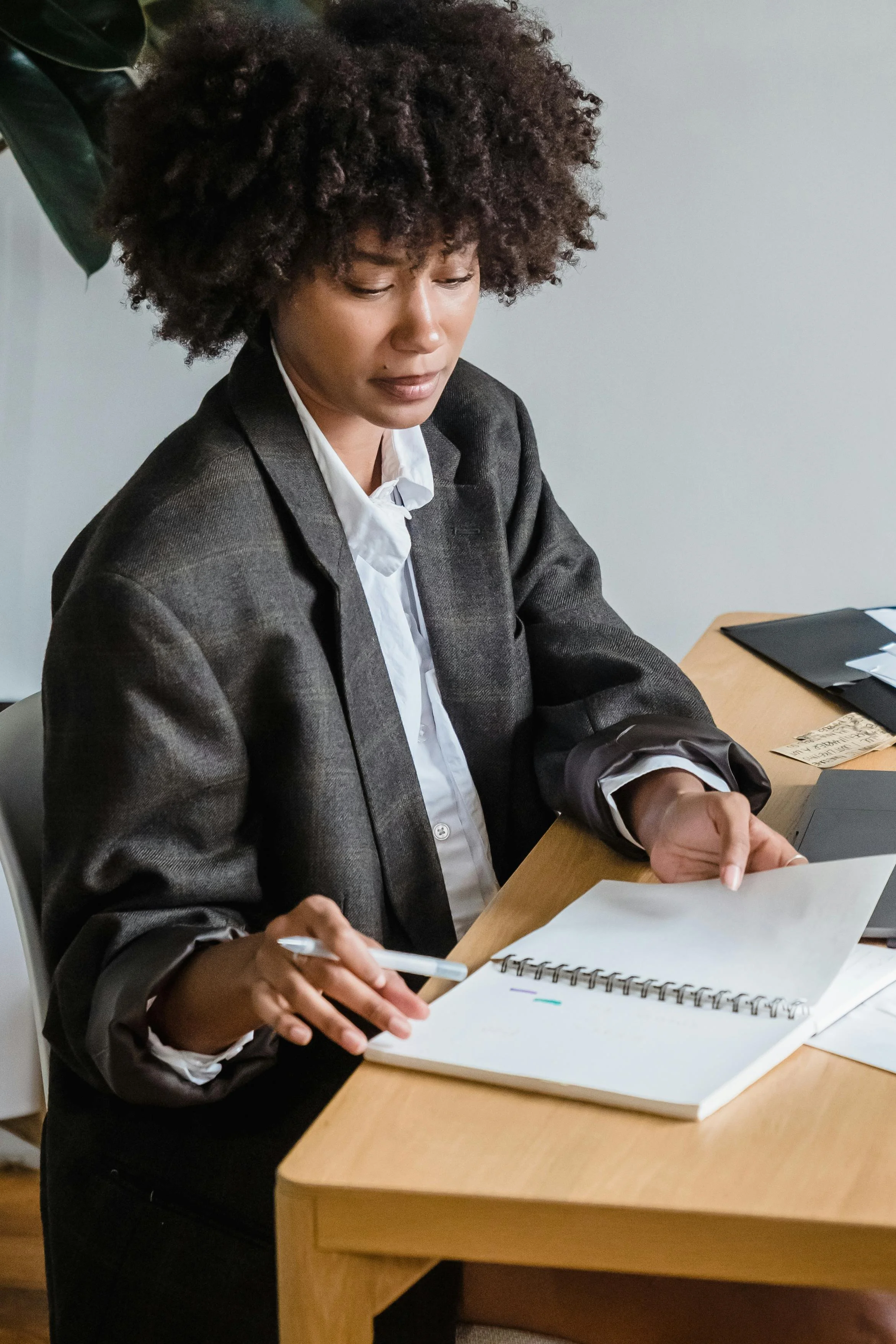 A woman with curly hair wearing a gray blazer and white shirt, sitting at a wooden desk, reviewing a sheet of paper and holding a pen. There is a black folder and a notebook on the desk, with a gray wall in the background.