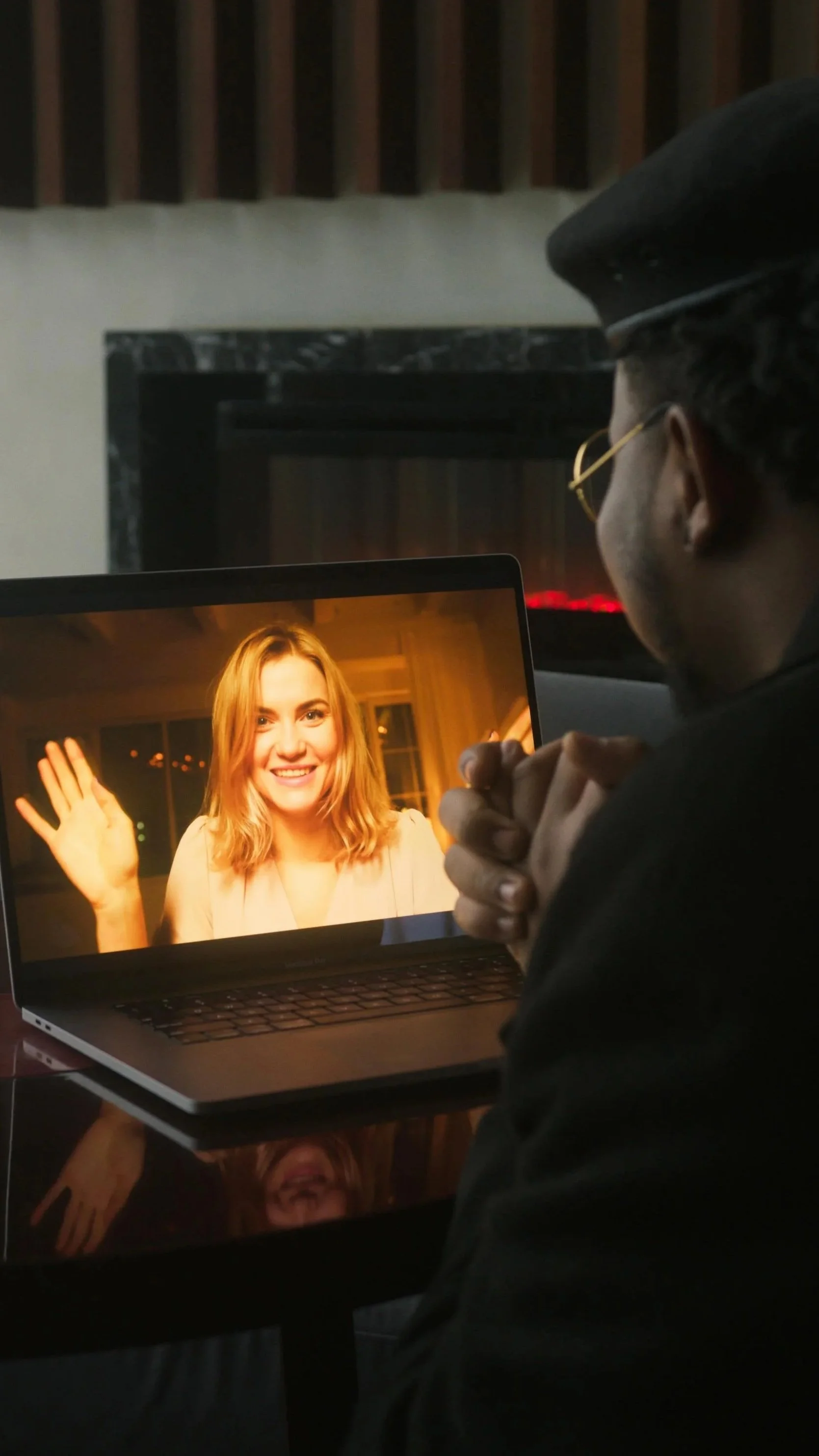 Person wearing a dark hat and glasses looking at a laptop screen that shows a woman with blonde hair waving and smiling.