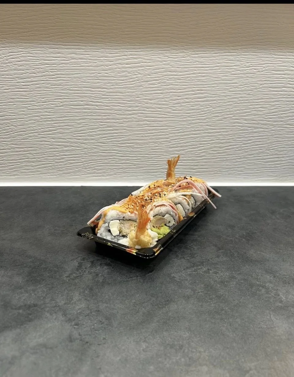 A black tray of assorted sushi rolls with shrimp tempura, avocado, and crab, garnished with sesame seeds and pink pickled ginger, placed on a dark gray surface against a beige wall with textured horizontal lines.