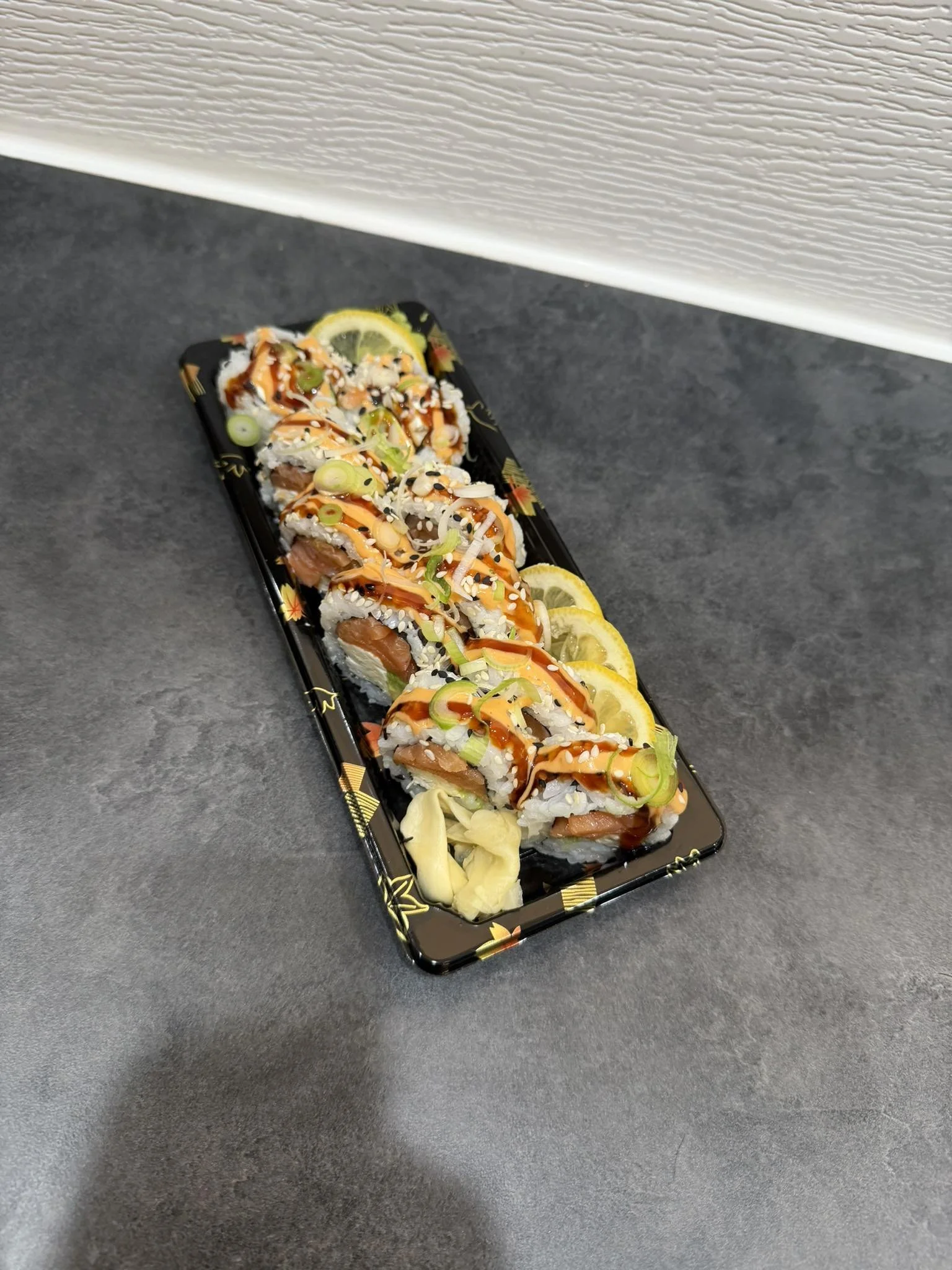 Sushi rolls arranged on a black tray with lemon slices and pickled ginger, garnished with sauces and sesame seeds.