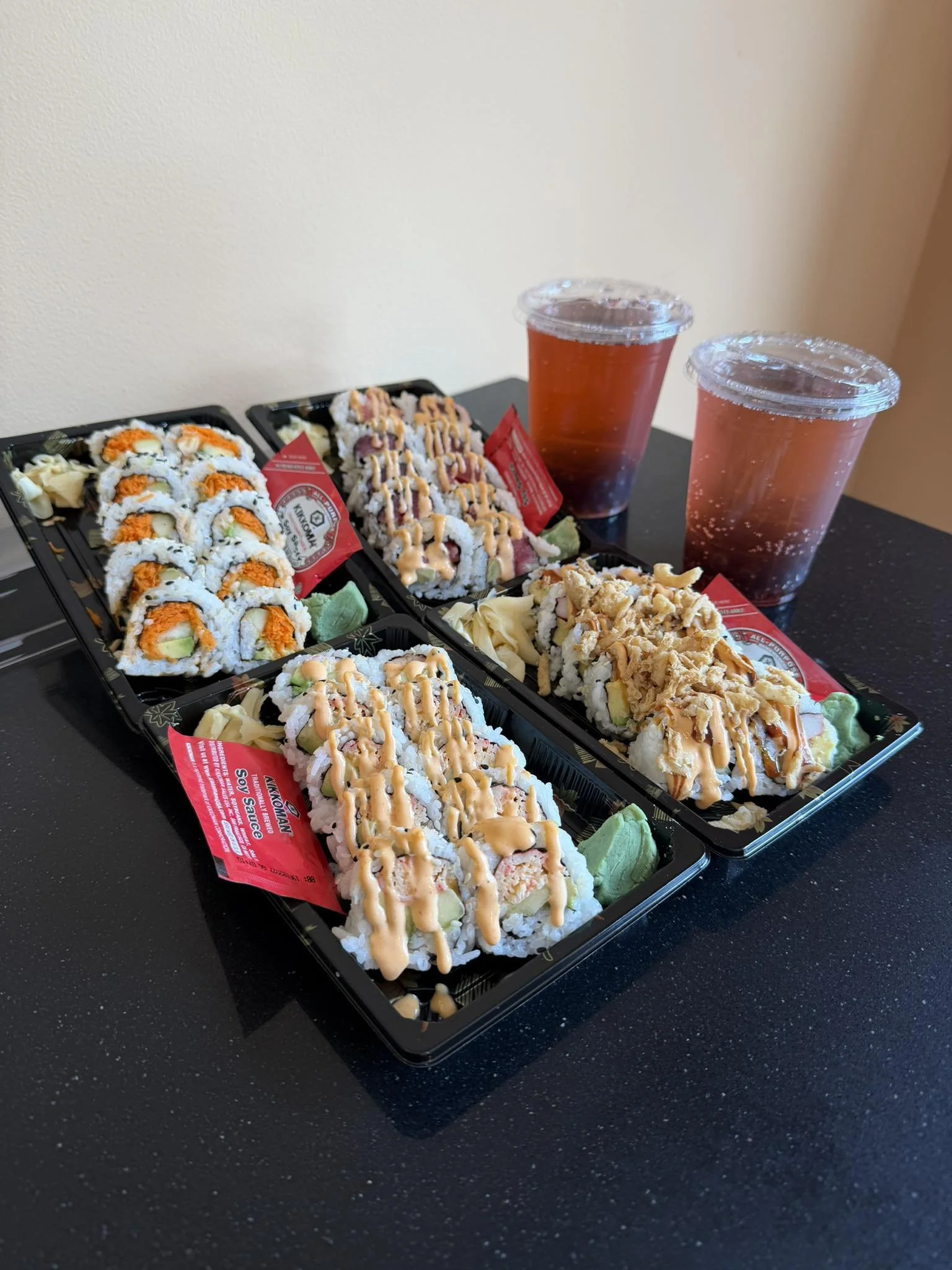 Two black plastic sushi trays filled with assorted sushi rolls topped with sauces, accompanied by wasabi and pickled ginger, with two cups of soda in the background on a black countertop.
