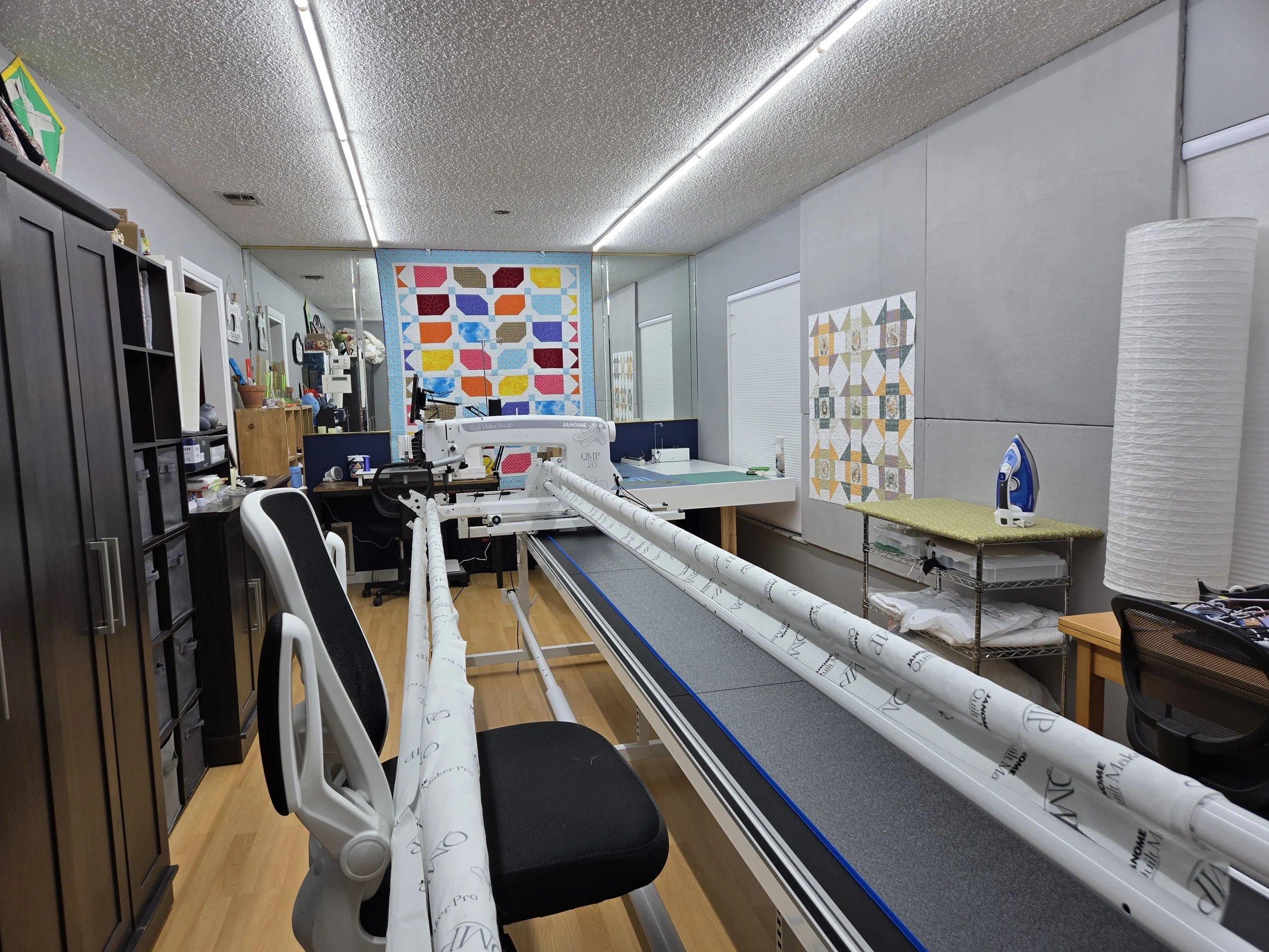 A sewing or quilting workspace with fabric rolls, a sewing machine, and quilted fabric pieces hanging on the wall.