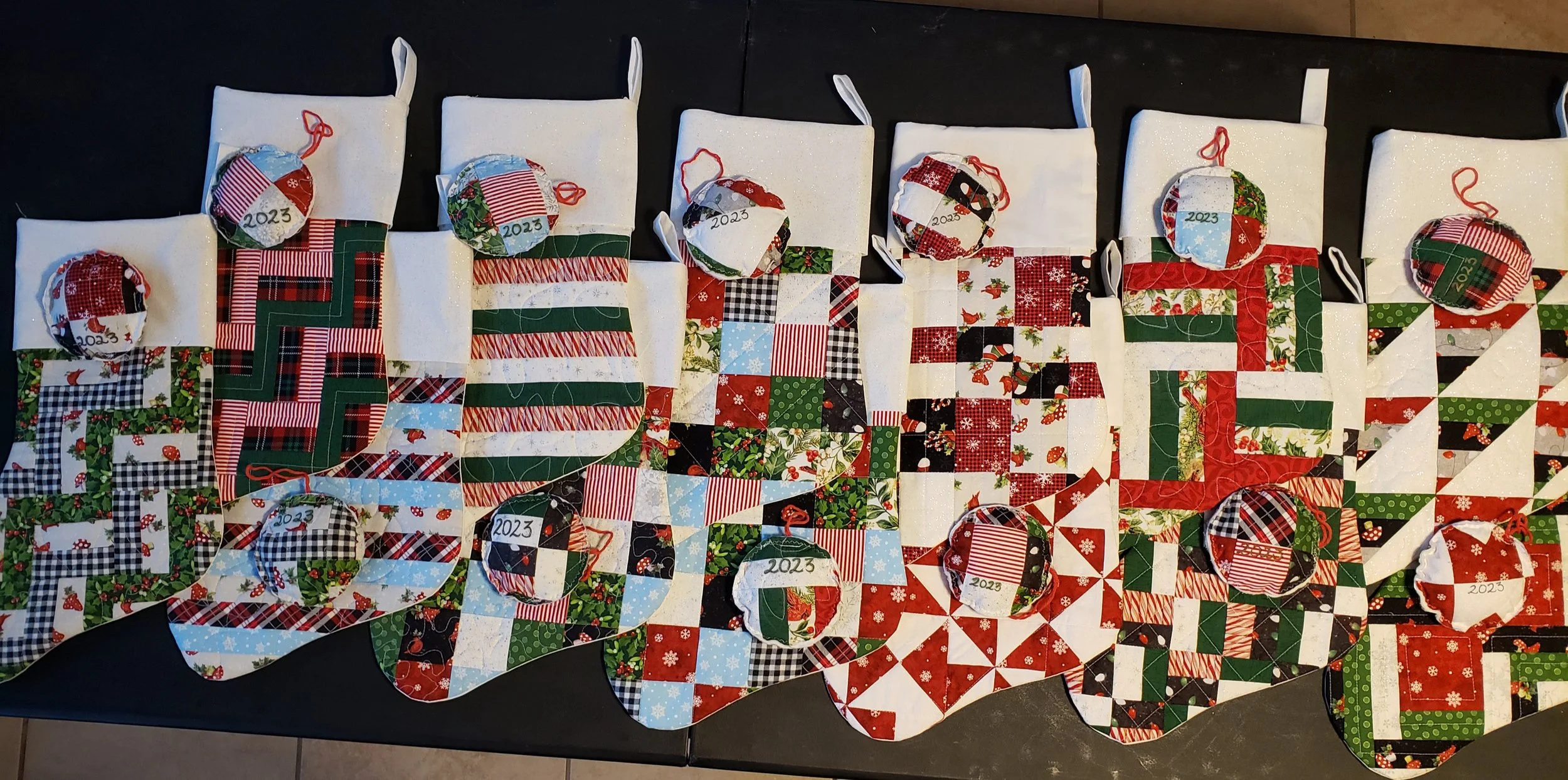A Christmas quilt with each square decorated with holiday-themed fabrics, and small Christmas stockings with red ribbons and '2023' tags on top, placed on a dark surface.