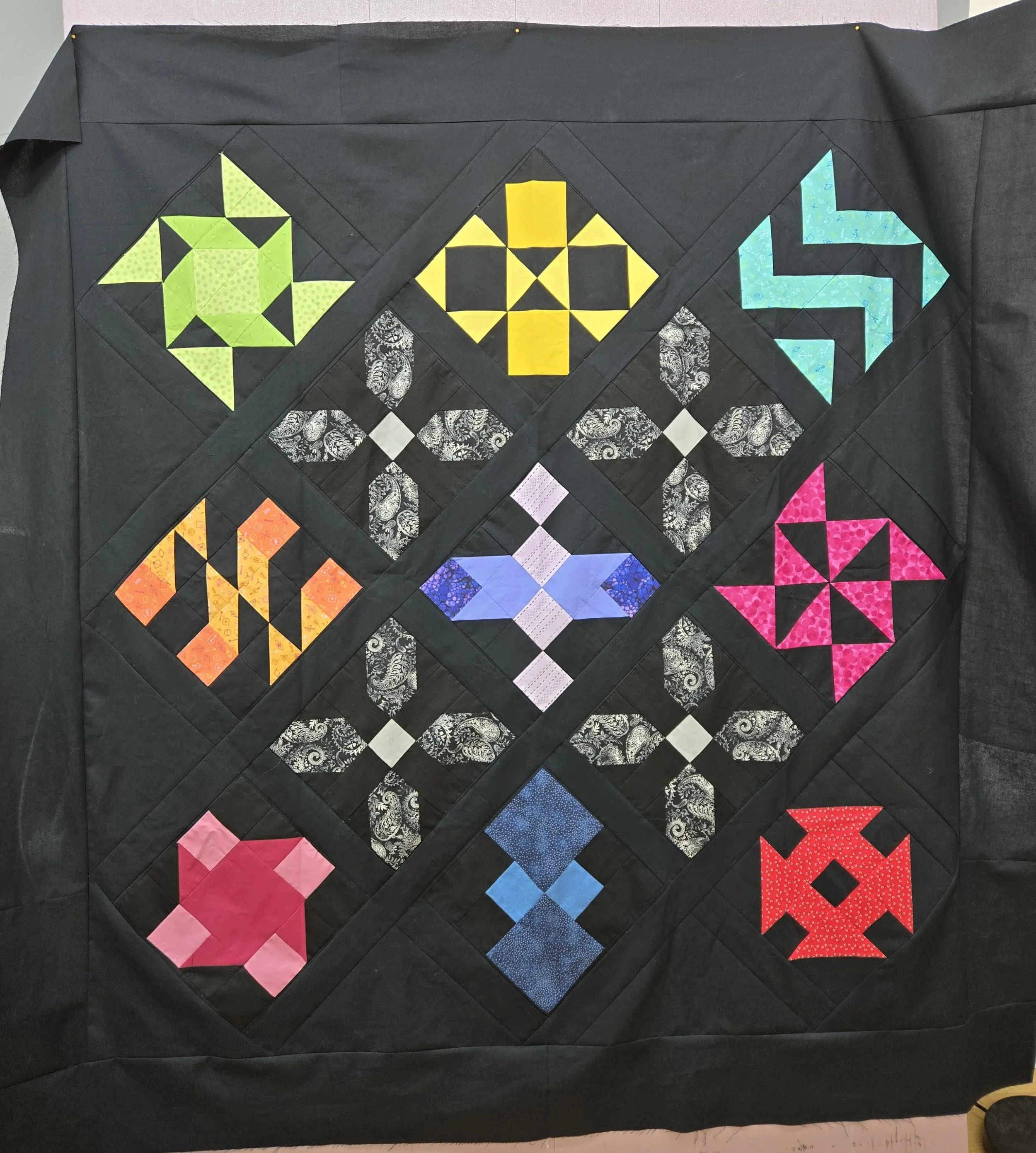 A quilt with a diamond shape pattern on a black background. The quilt features colorful star and geometric designs in various colors including yellow, teal, pink, orange, blue, and black. The patterns incorporate different fabrics and textures, creating a vibrant mosaic.