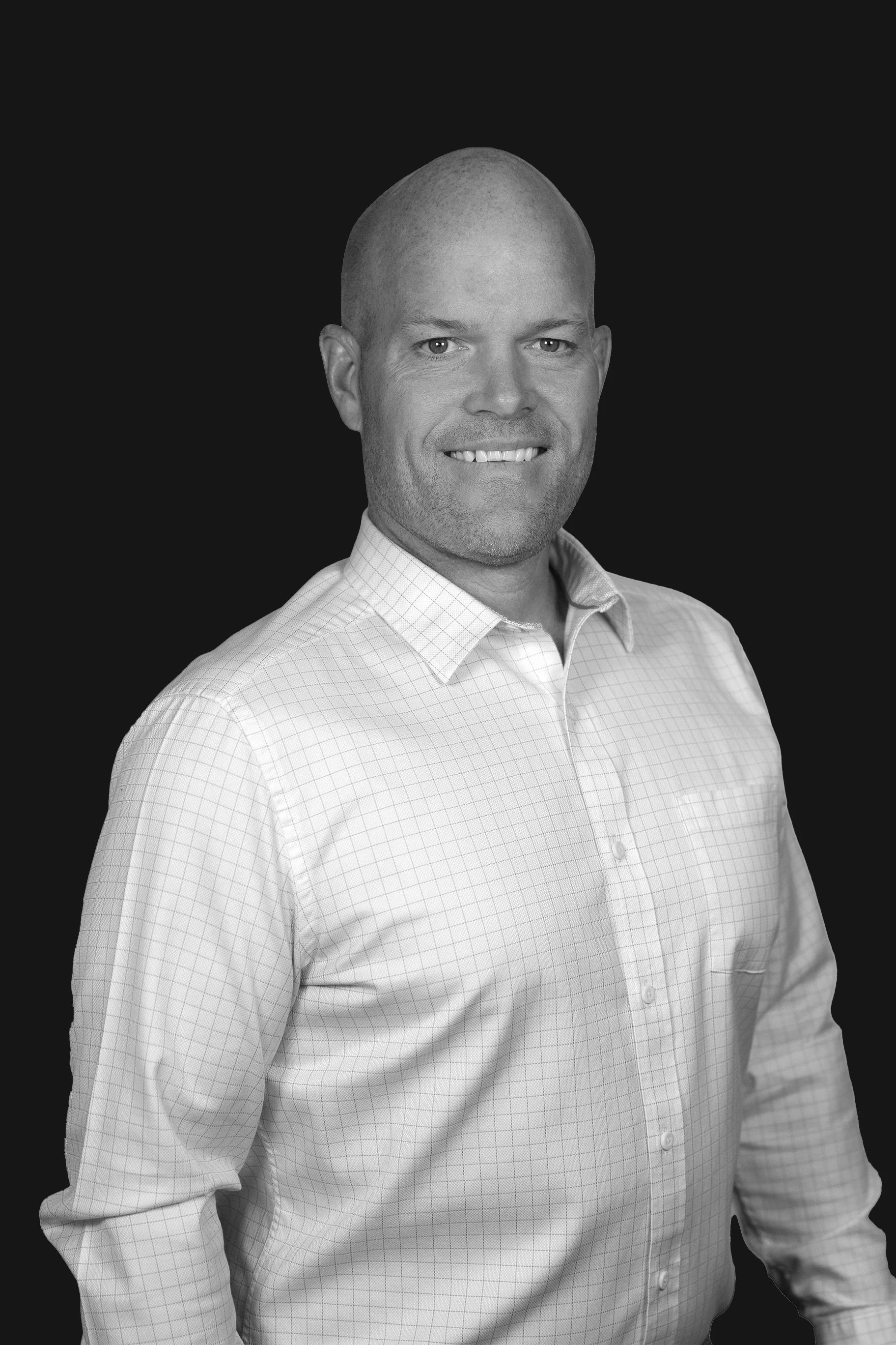 A black and white photograph of a smiling man with a shaved head, wearing a light-colored checkered dress shirt against a dark background.