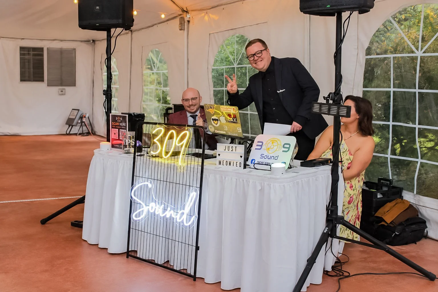 People at a DJ or radio station table inside a white tent. Three individuals are present: a woman in a floral dress, a man in a suit with glasses, and another man standing in a suit. The table has neon signs, laptops, and equipment. The woman is sitting, the man with glasses is sitting, and the standing man is smiling and making a peace sign. There are decorative lights, and greenery is visible through the tent windows.
