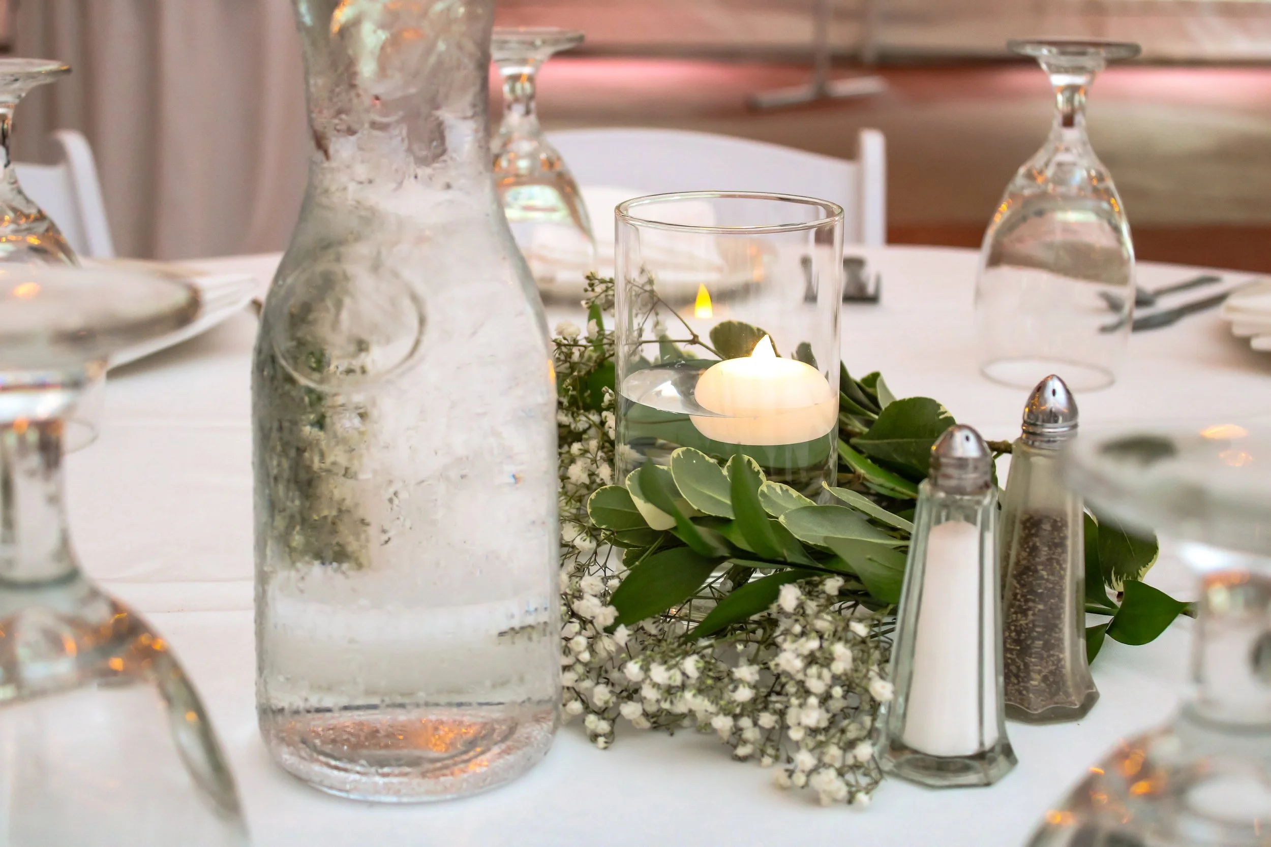 A table decorated with glass vases, a floating candle, greenery, and small white flowers, set for a special meal or celebration.