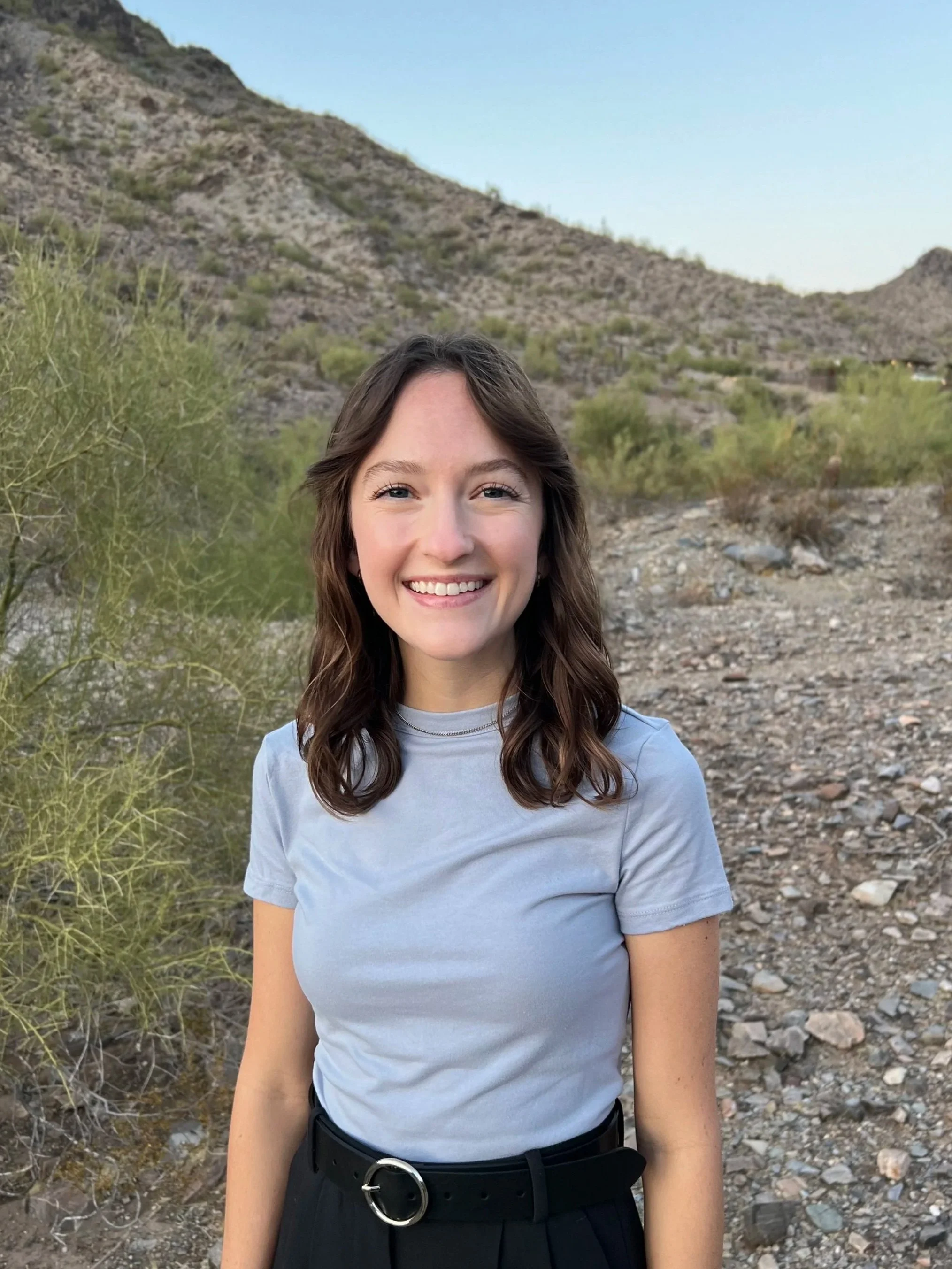 A young woman with long brown hair smiling outdoors in a desert landscape with mountains and sparse desert plants in the background.