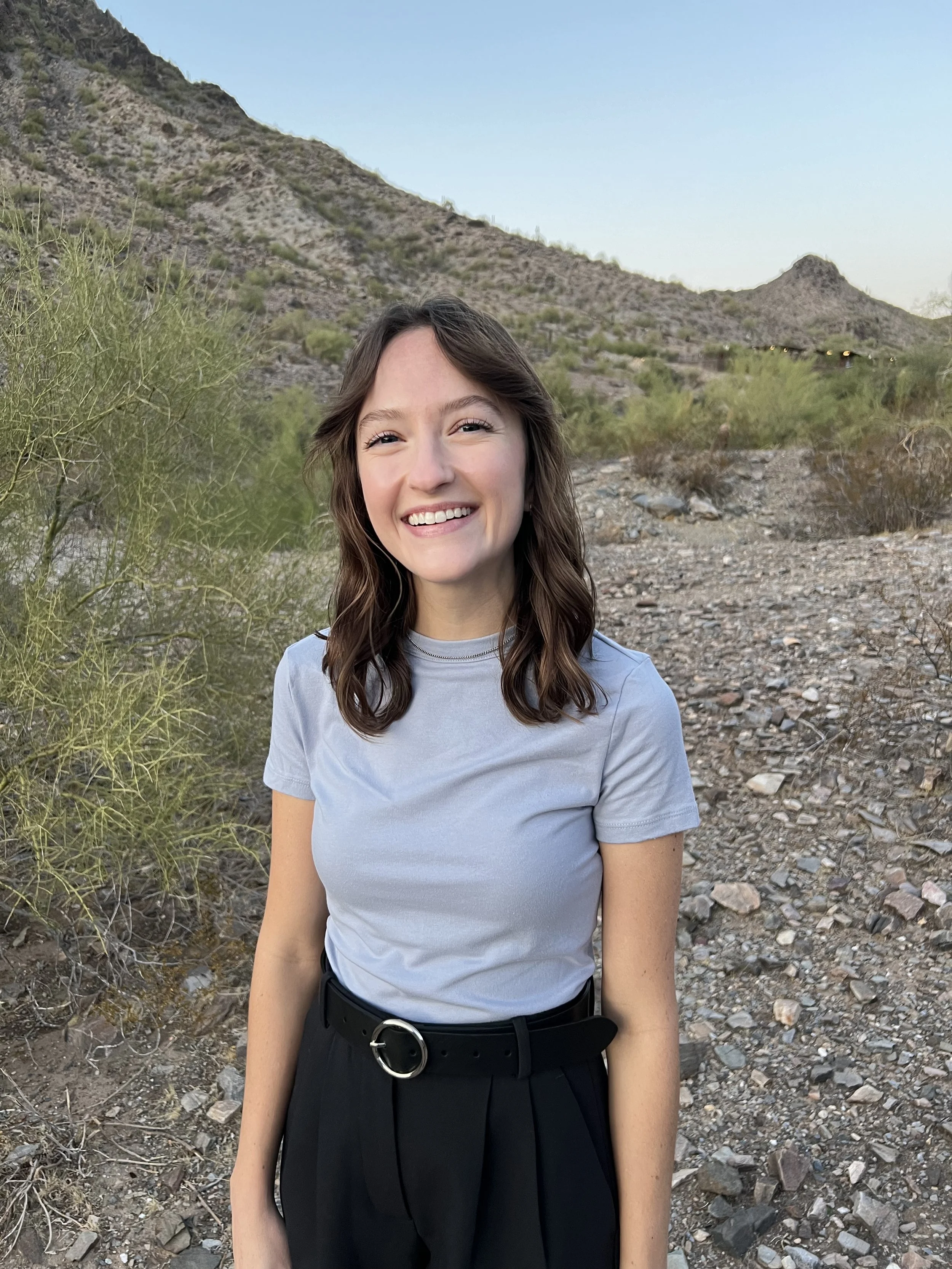 A woman with brown wavy hair smiling outdoors with desert mountains and sparse vegetation in the background.