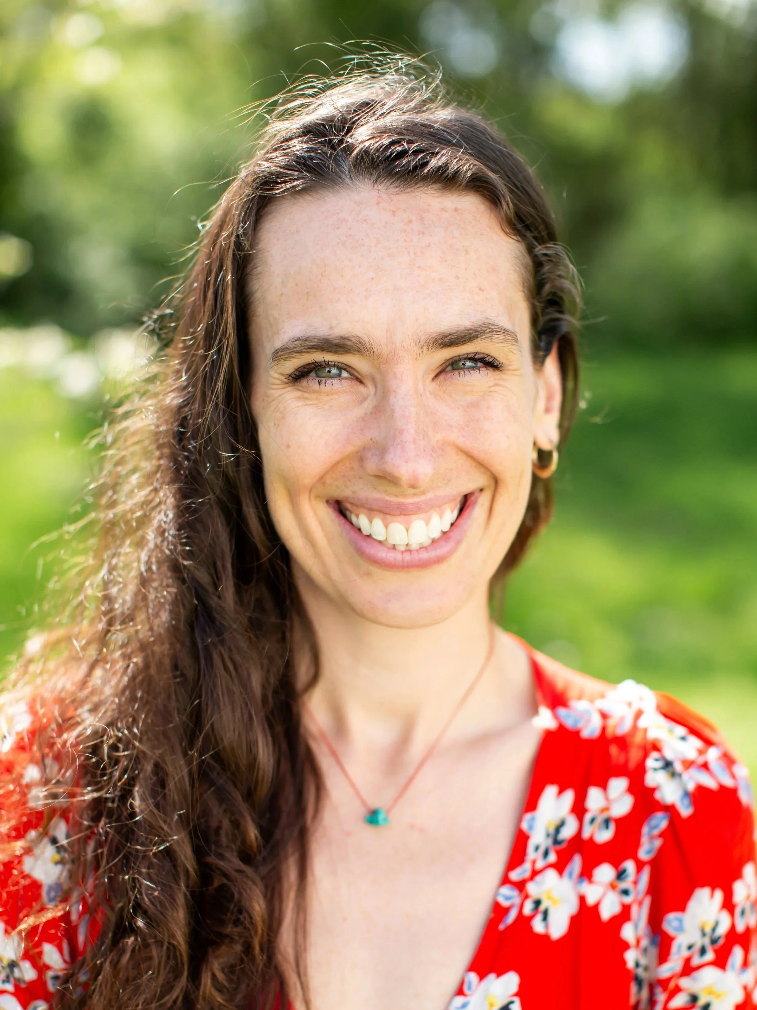 Close-up of a woman smiling outdoors with green blurred background, wearing a red floral dress and a turquoise necklace.