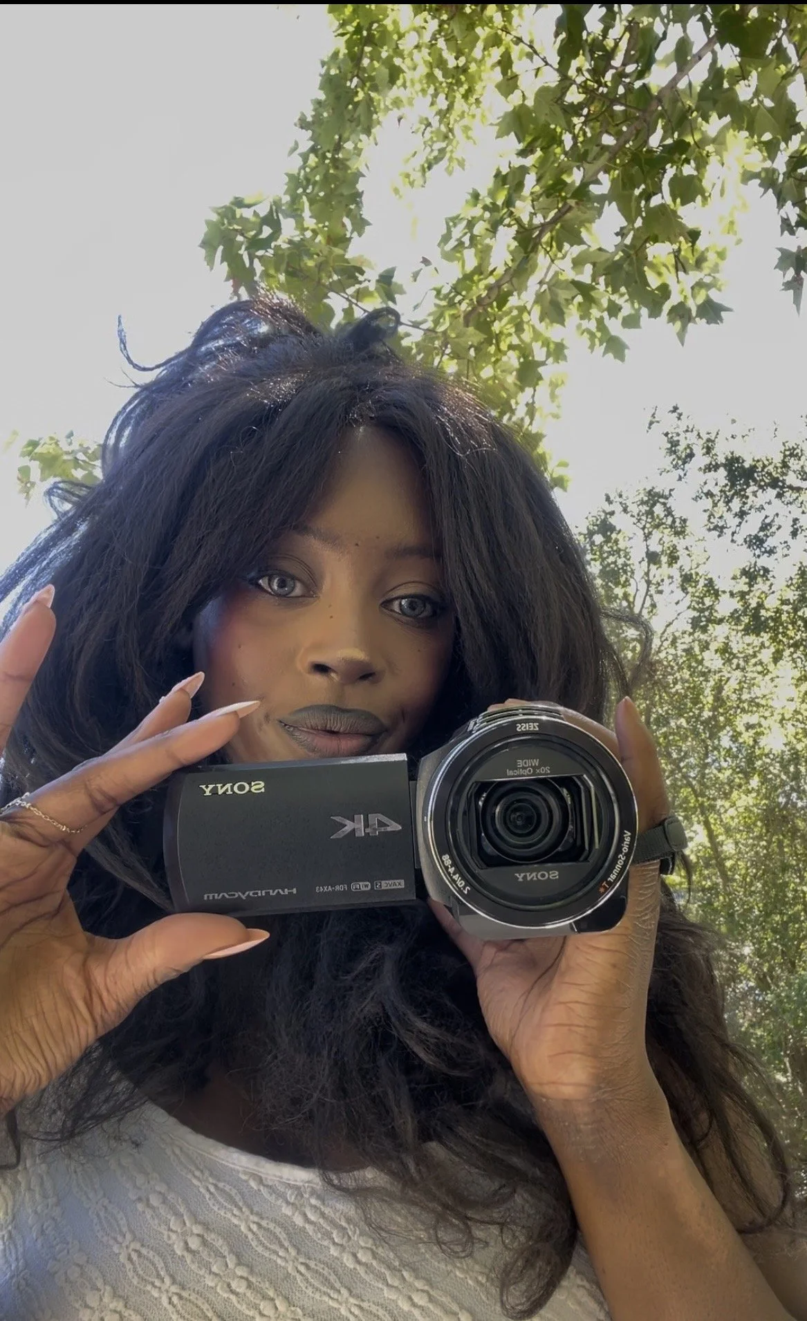 A woman with dark hair and light eye color taking a selfie outdoors with a camera, surrounded by green trees and sunlight.