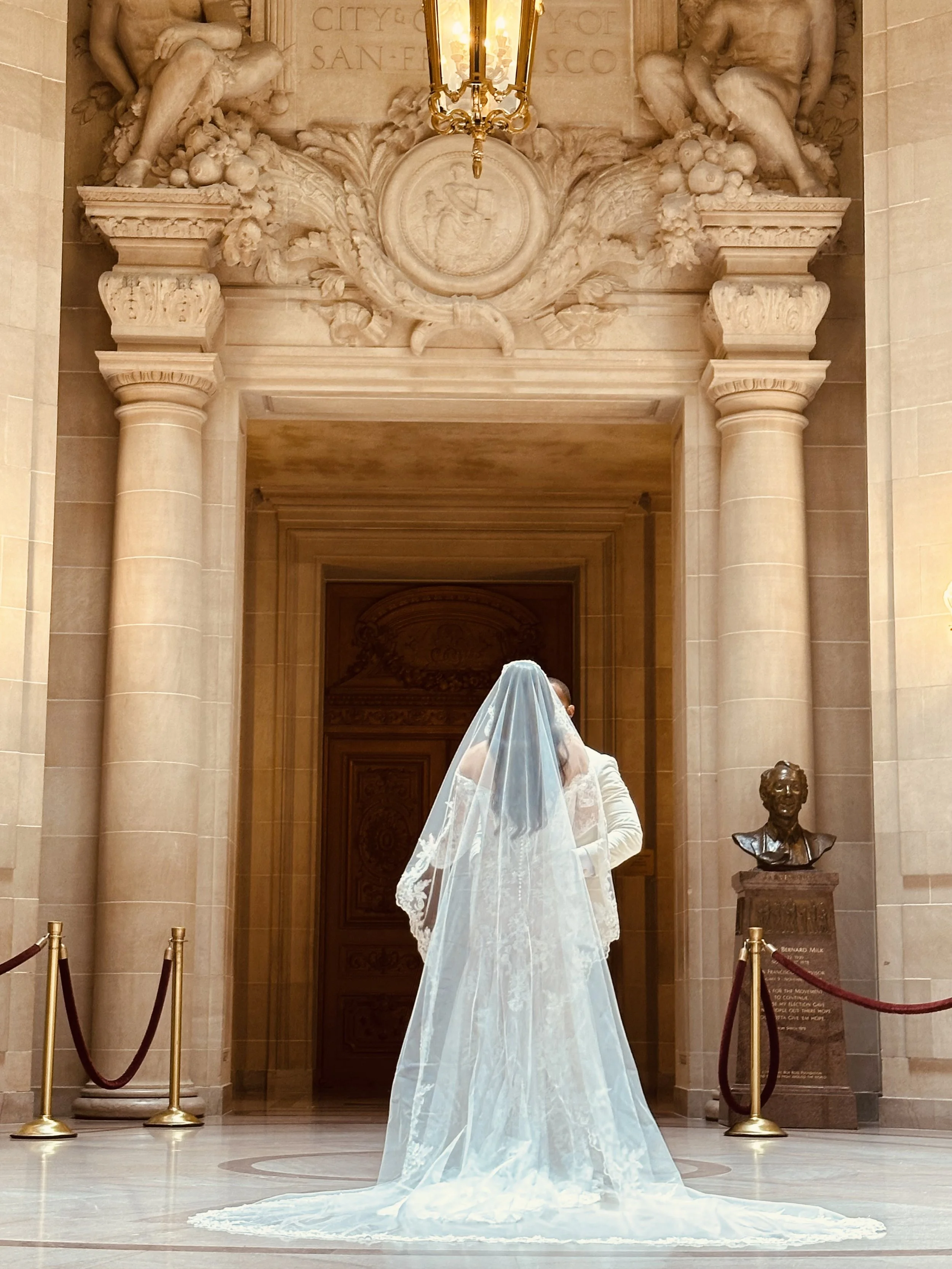 A person dressed in a wedding gown with a long veil standing in a historic building lobby with tall columns, ornate carvings, and a bust sculpture.
