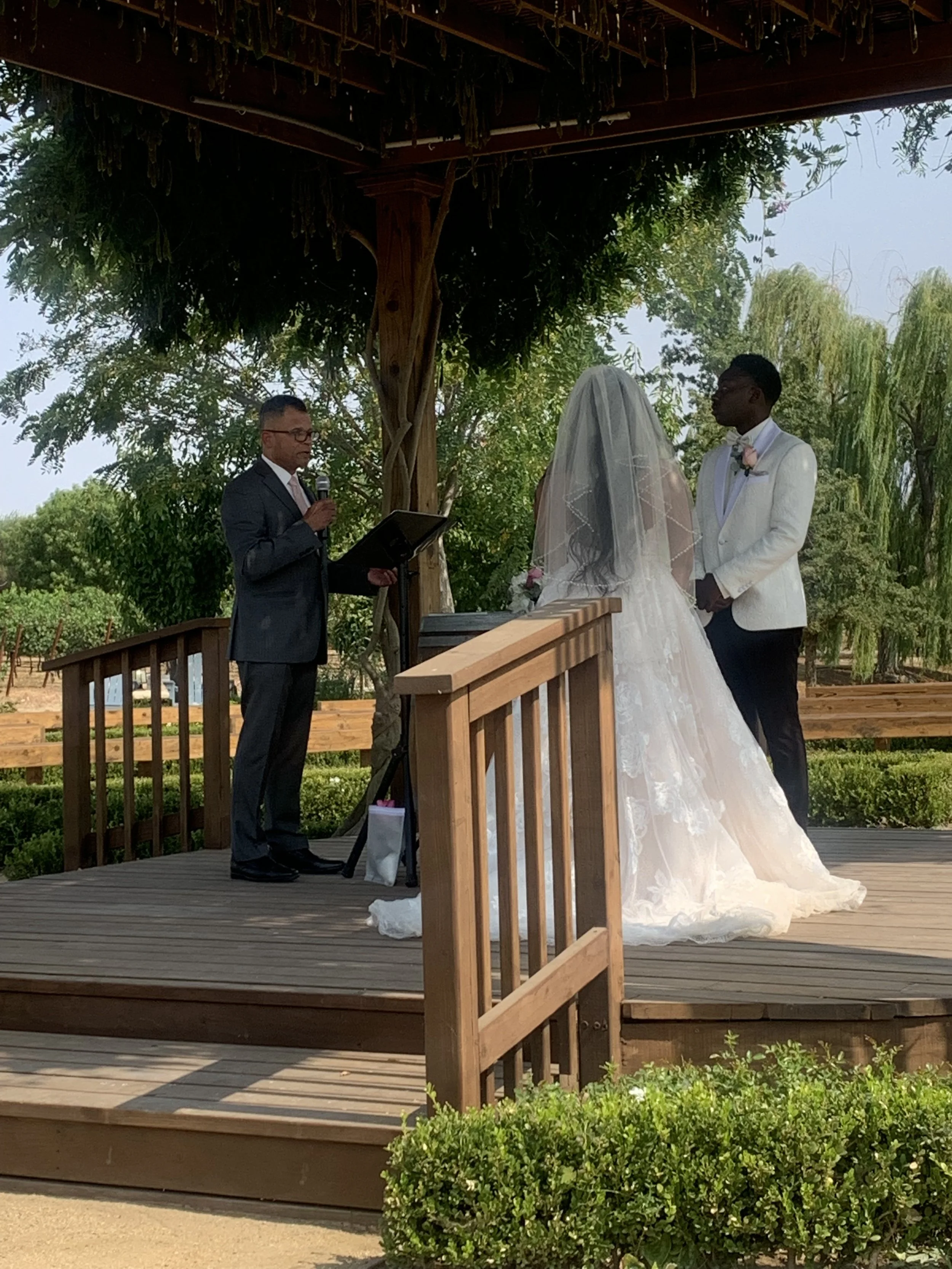 A wedding ceremony taking place outdoors under a canopy of trees. A man in a dark suit is speaking into a microphone, while a bride in a white wedding gown and veil, and a groom in a white tuxedo with a pink boutonniere, stand beside him. The ceremony is on a wooden stage with a railing, surrounded by greenery and trees.