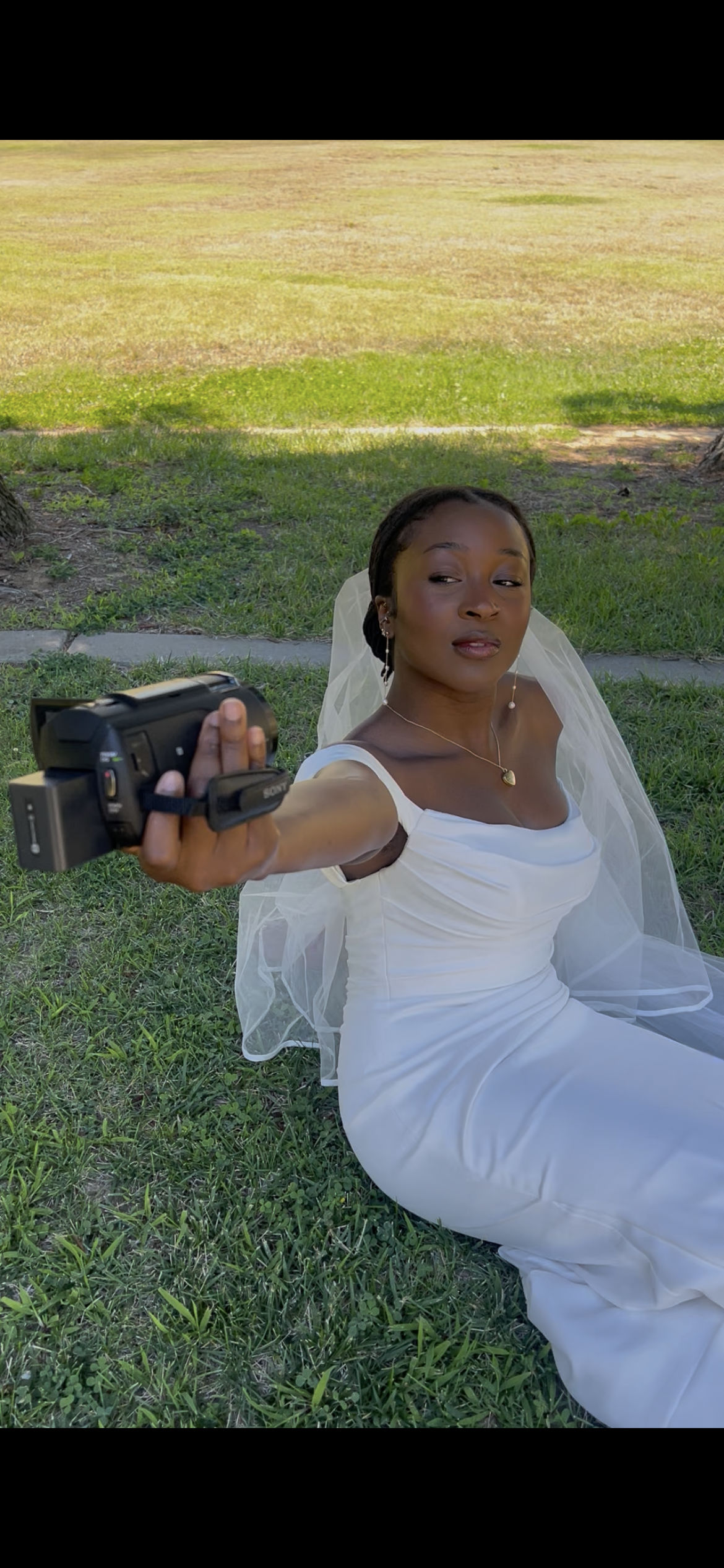 A woman in a white dress, sitting on grass outdoors, wearing a veil and jewelry, taking a selfie with a video camera held out in front of her.