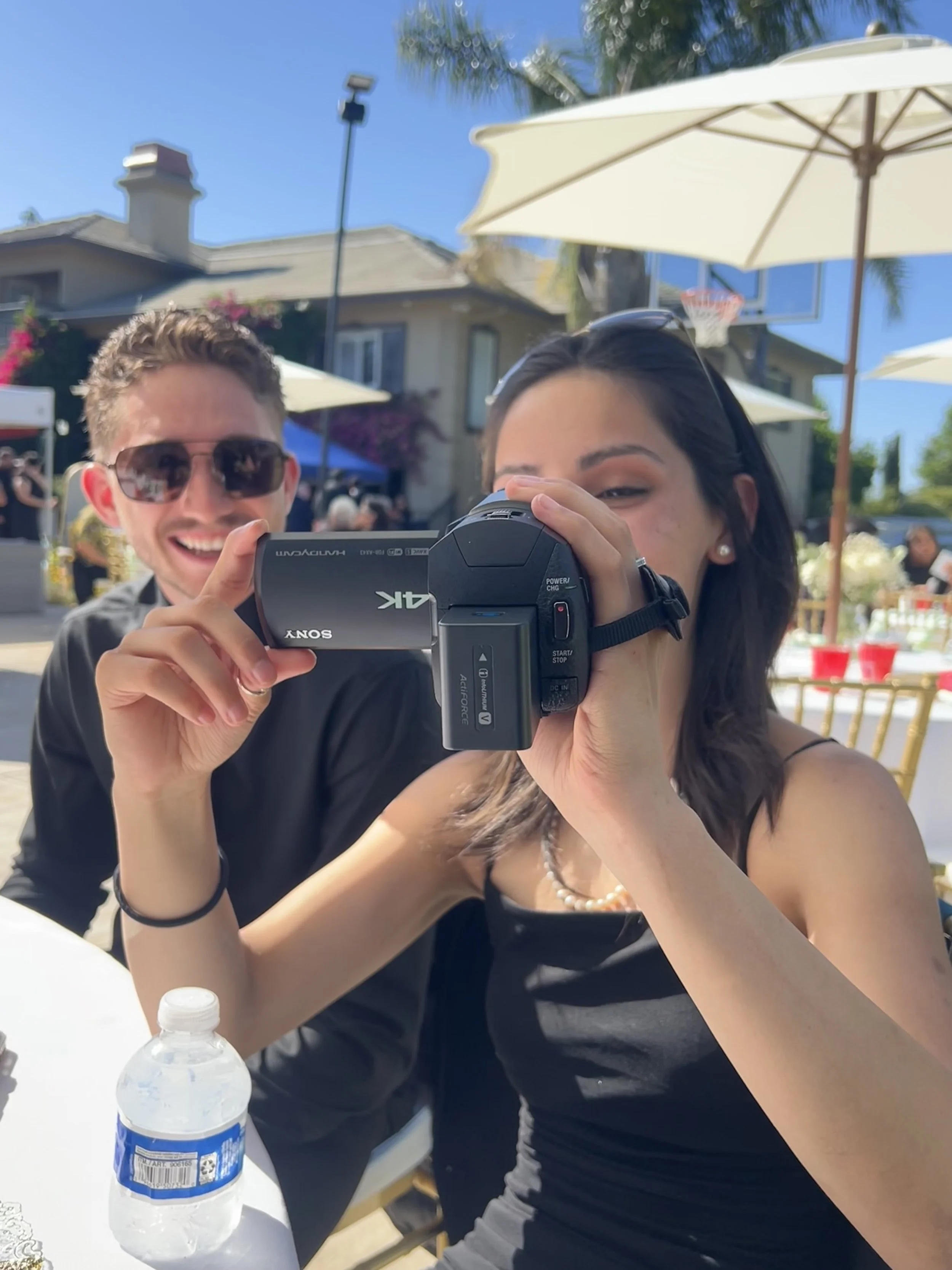 A woman with dark hair and pearl earrings taking a photo with a camcorder at an outdoor gathering. A man with sunglasses and a black jacket is smiling in the background. The scene is bright with blue sky, umbrellas, a house, and other people in the background.