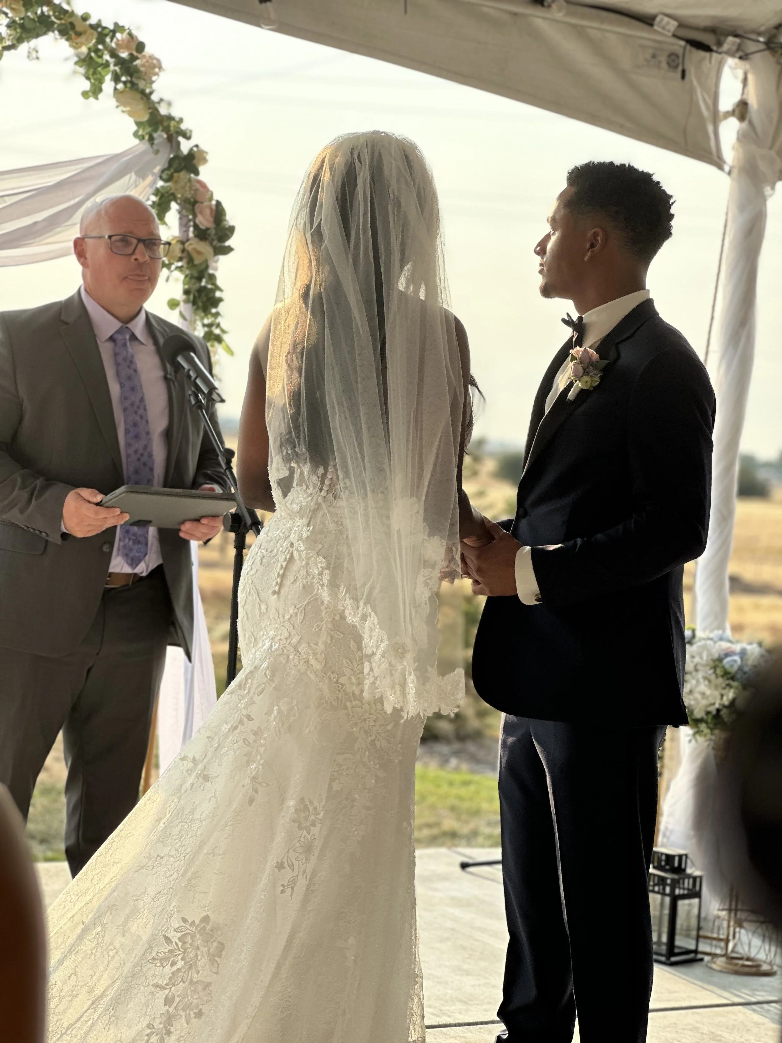 A bride and groom stand facing each other during their wedding ceremony, holding hands under a decorated outdoor canopy, with an officiant to the left.