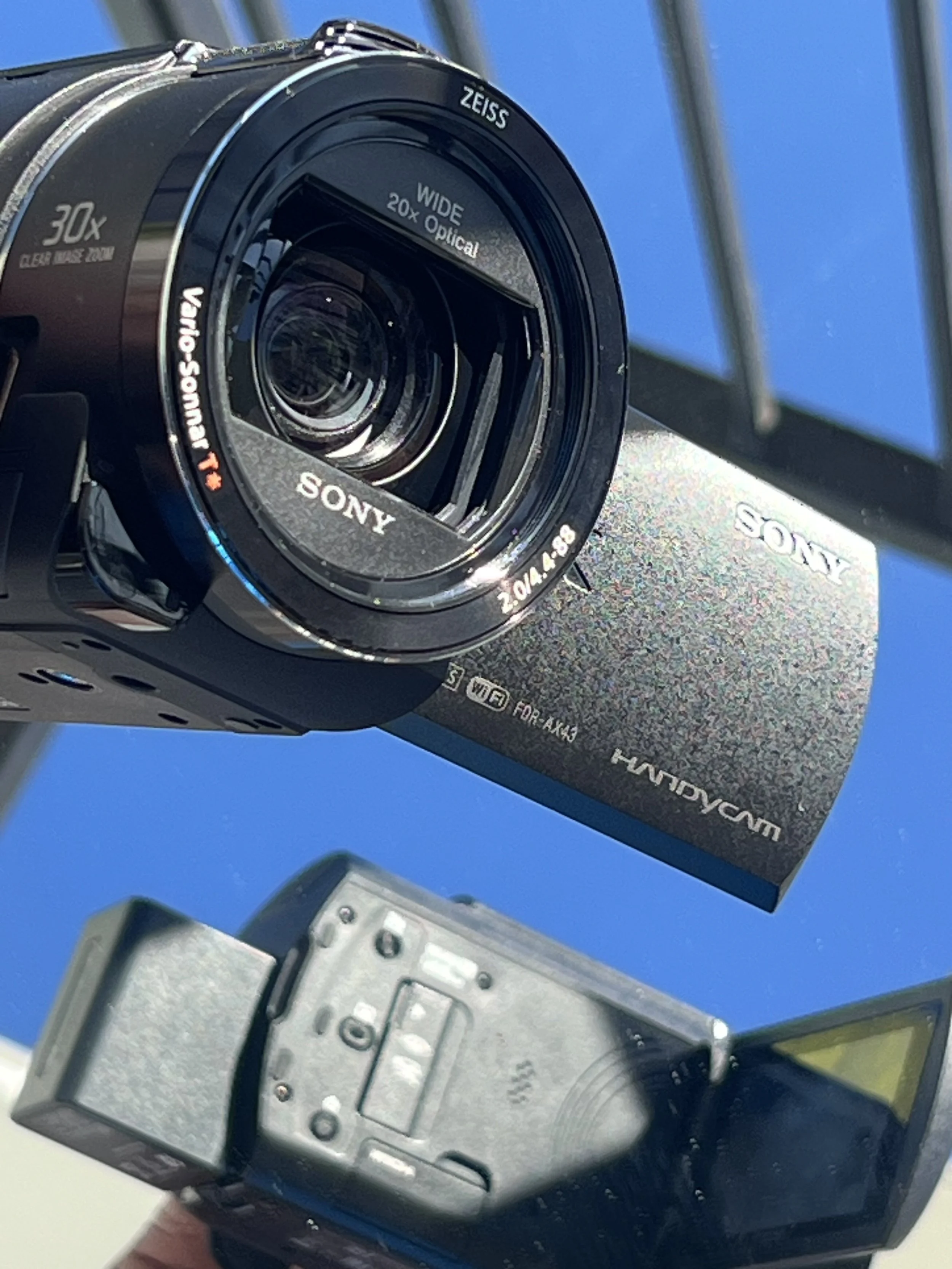 Close-up of a Sony camera lens with 30x optical zoom, mounted in front of a city skyline with buildings and a clear blue sky.