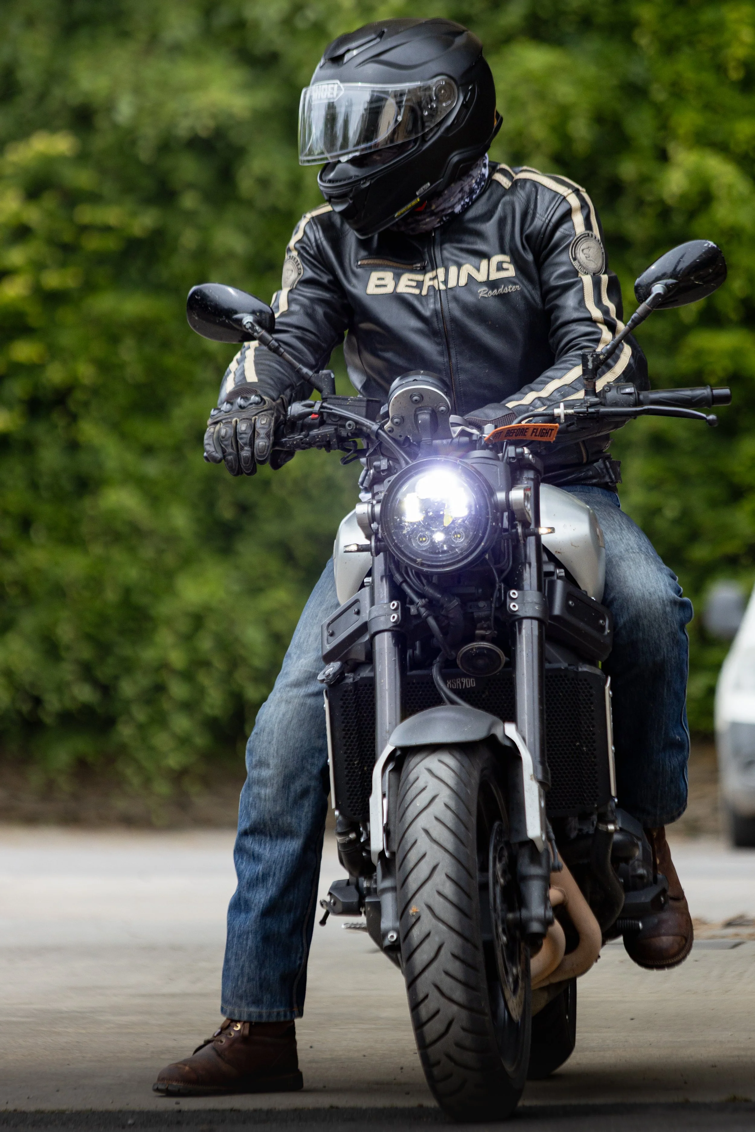 A person wearing a black full-face helmet and a black leather jacket with beige stripes riding a motorcycle on a road with green trees in the background.