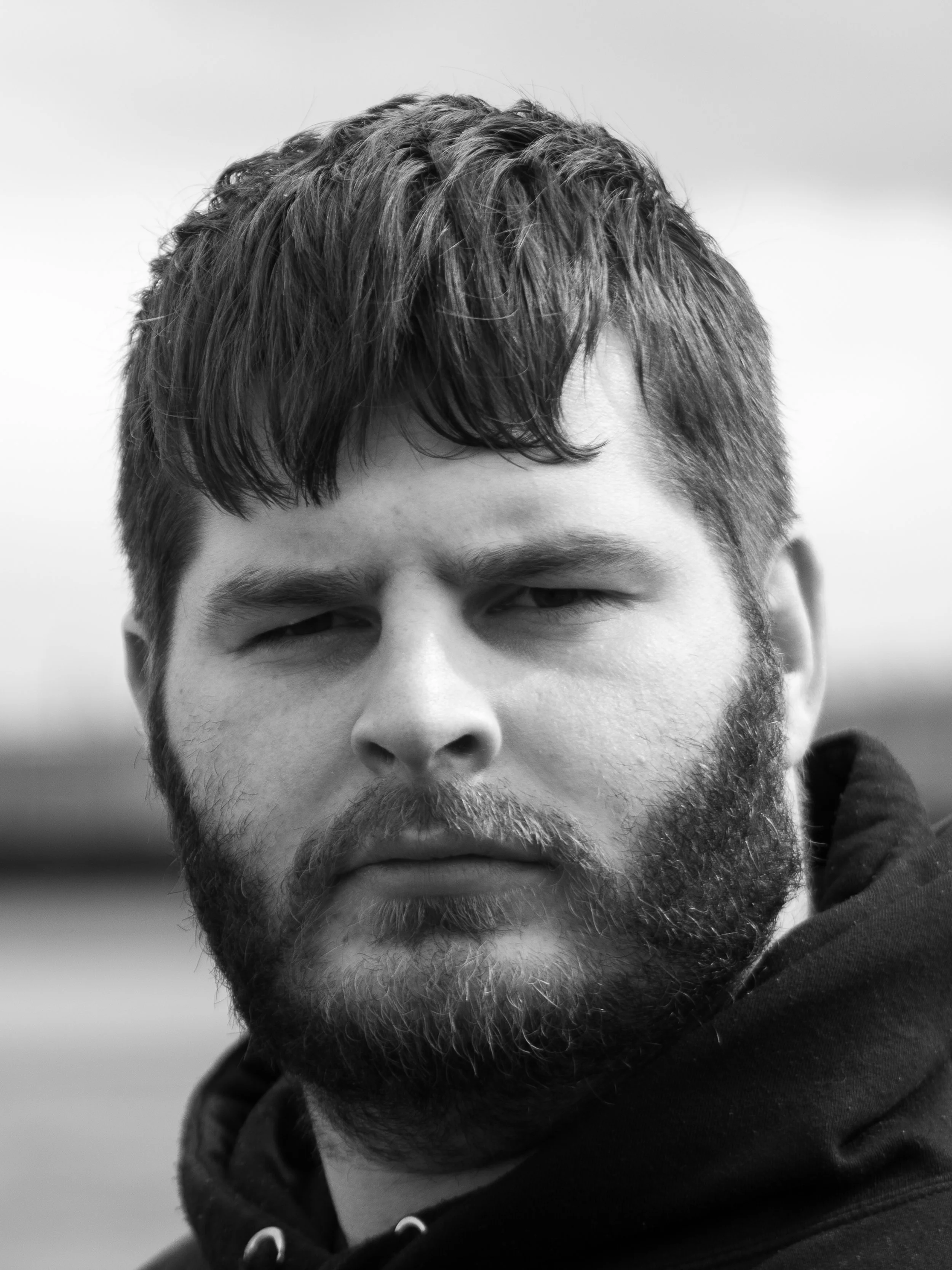 Close-up black and white photo of a young man with short, textured hair, a full beard, and mustache, wearing a hoodie.
