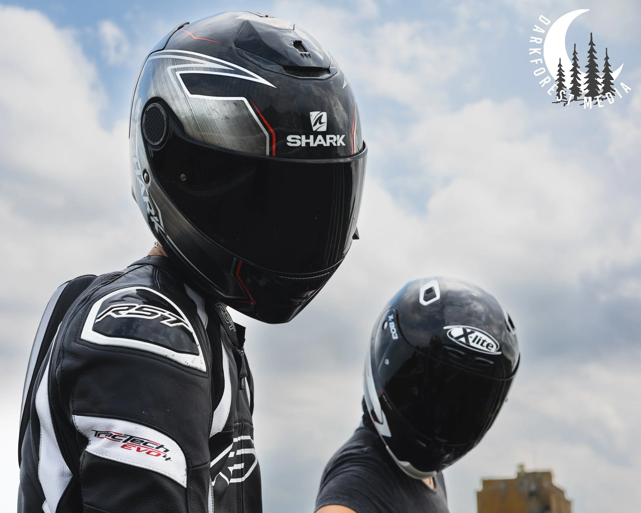 Two individuals wearing black full-face helmets and motorcycle gear standing outdoors against a partly cloudy sky.