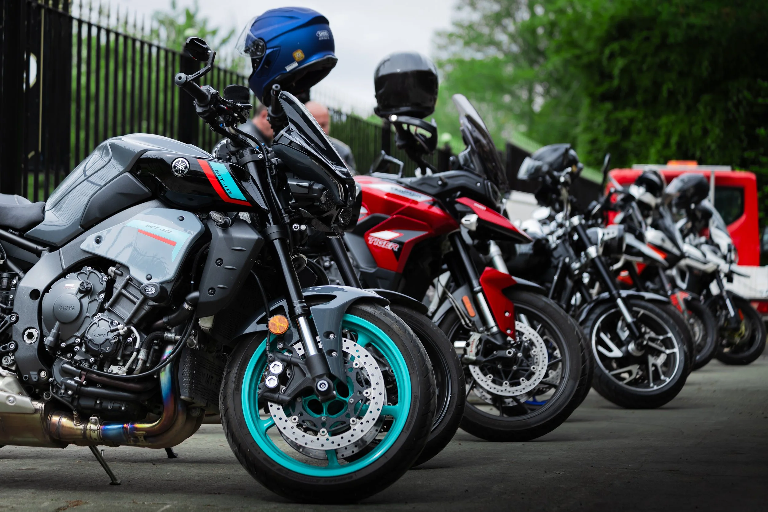 Multiple motorcycles parked in a row, with helmets placed on top of some of them, in an outdoor setting with trees and a fence in the background.