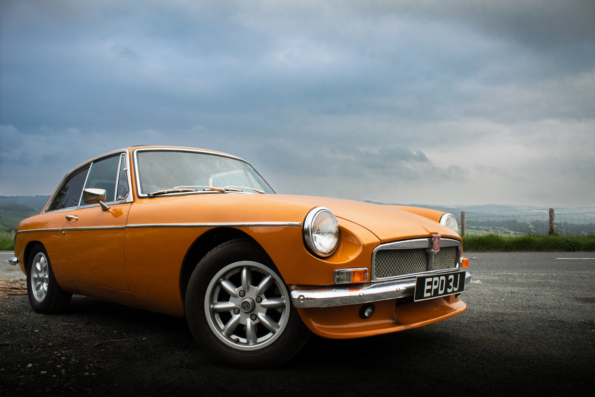 An orange vintage MG car parked on a roadside with a cloudy sky overhead, countryside in the background.