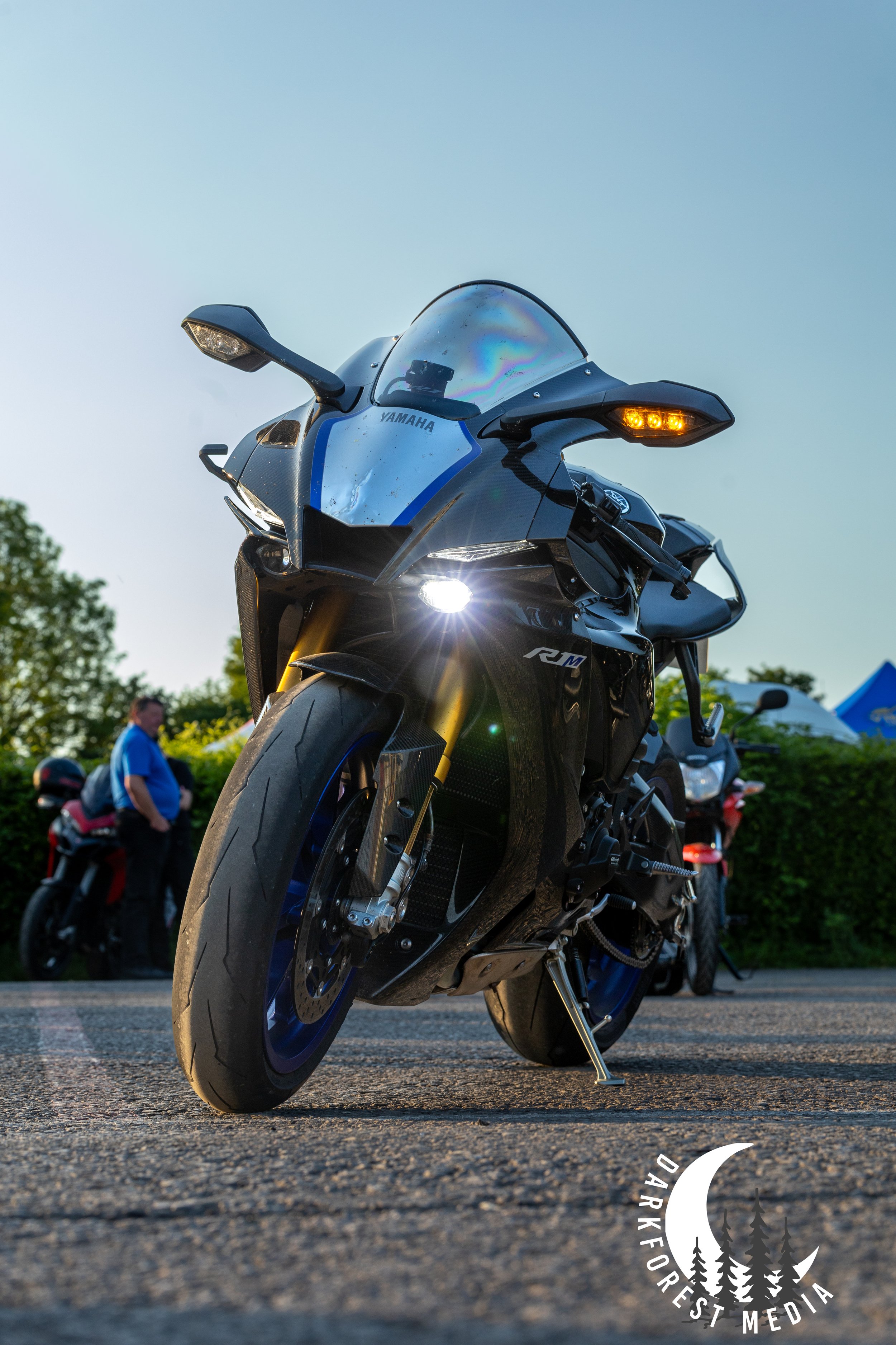 Close-up of a black Yamaha sport motorcycle parked on an asphalt surface, with blurred people and trees in the background during sunset.