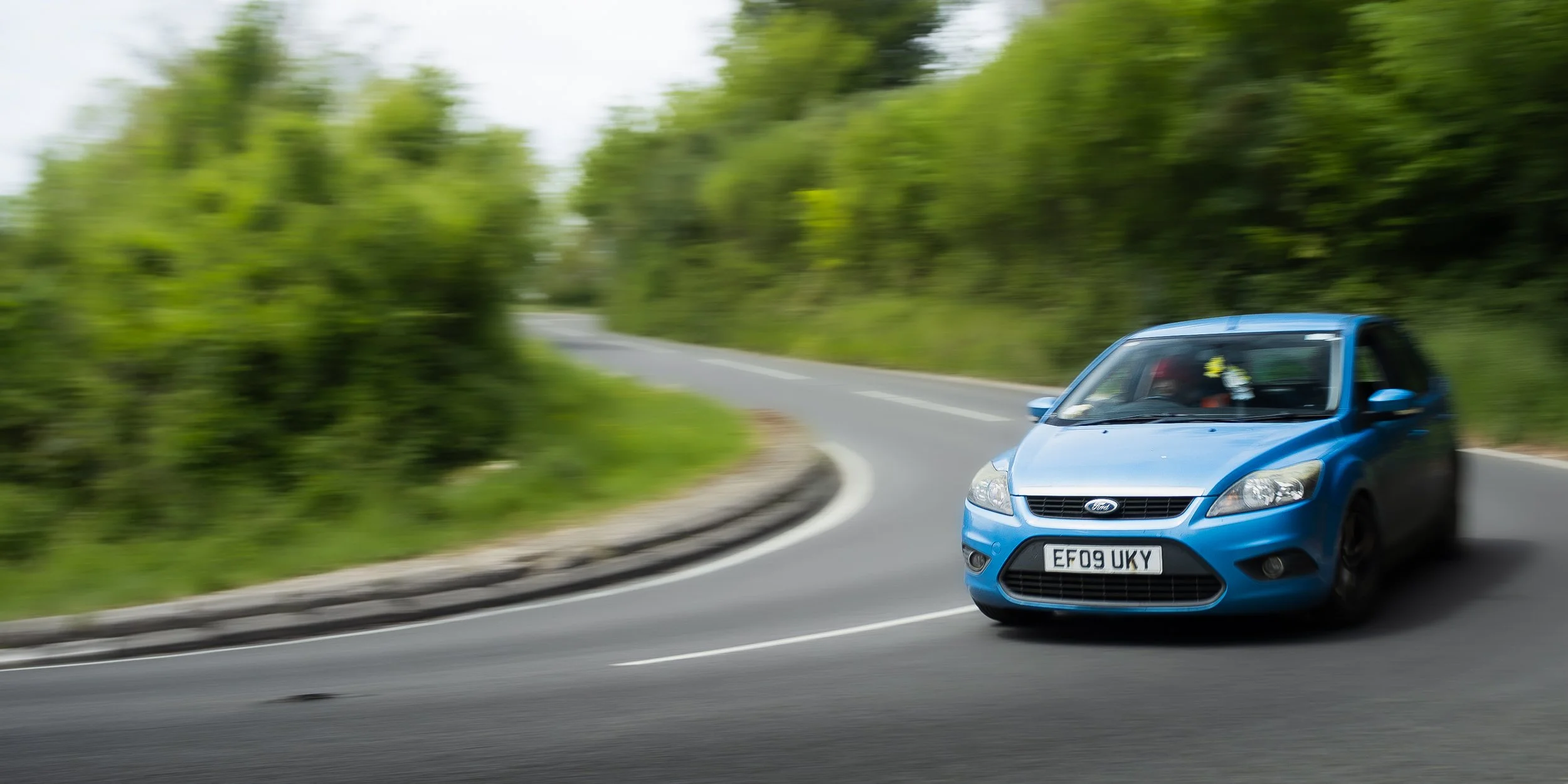 Blue Ford car driving on a winding road surrounded by green trees and foliage.
