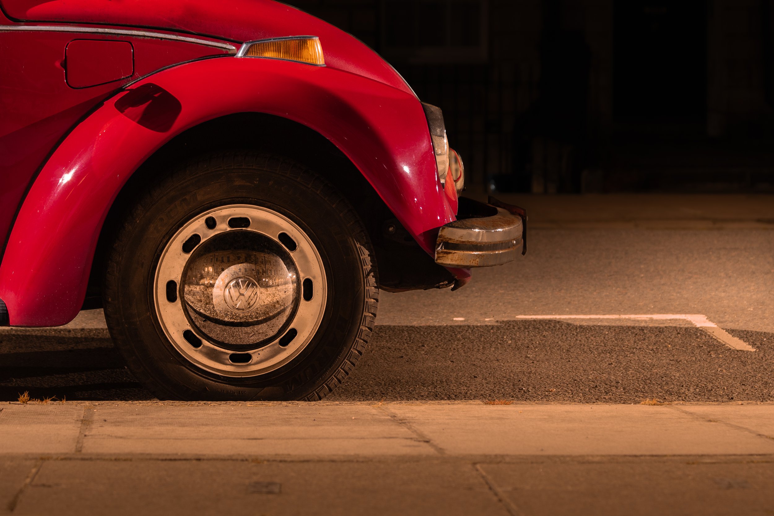 Close-up of the front portion of a vintage red Volkswagen Beetle parked on a street at night, showing the front wheel, bumper, and headlight area.