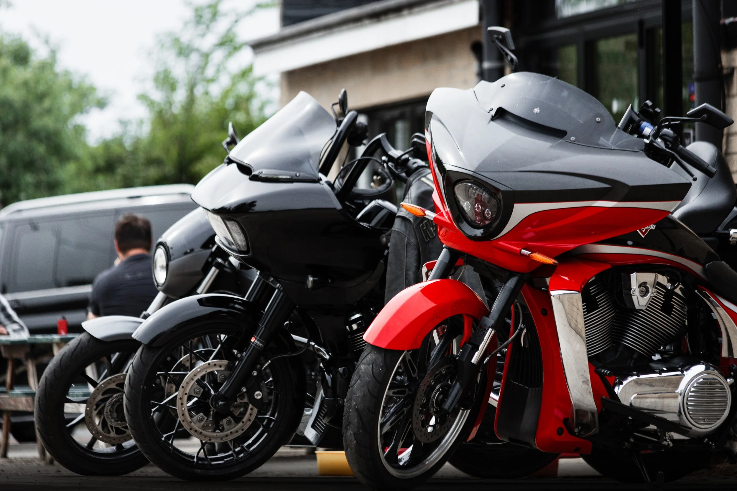 Several motorcycles parked in a lot with people nearby, some trees and a building in the background.
