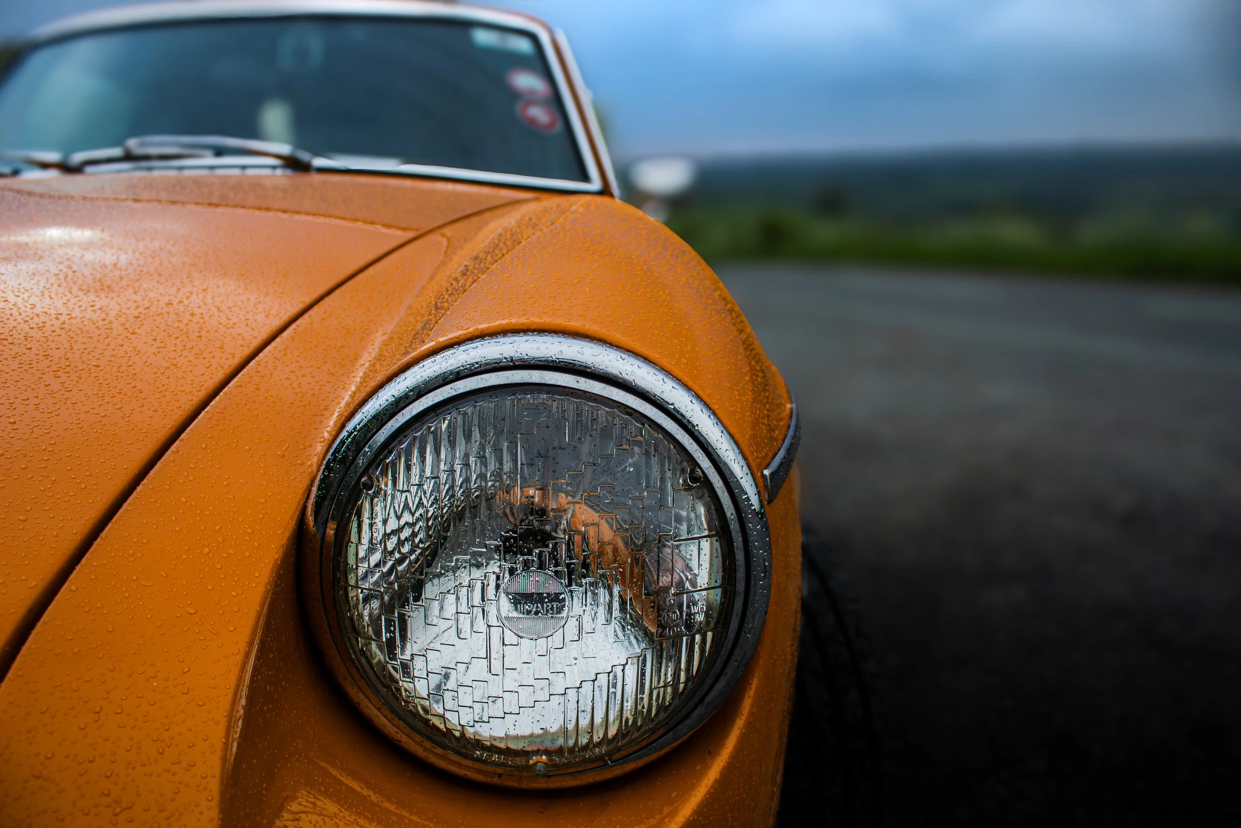 Close-up of a vintage orange car's headlight with raindrops on the hood, set against a blurred background of a roadside landscape.