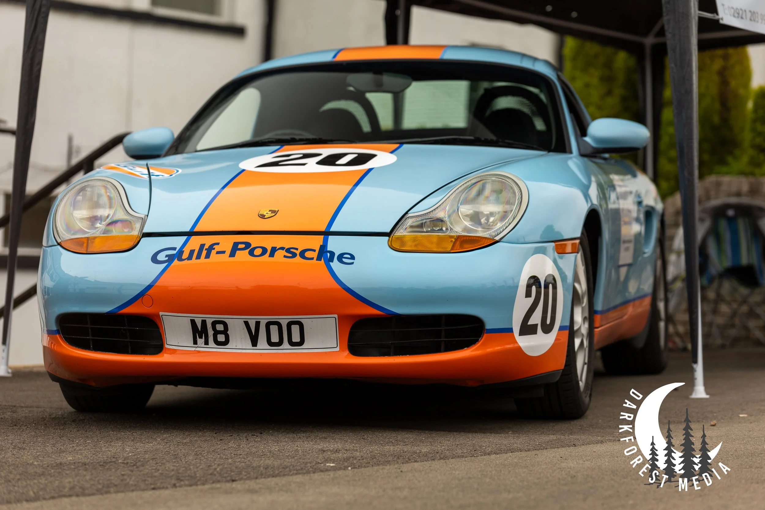 A vintage Porsche race car painted in light blue with orange racing stripes, the number 20 on the hood and side, and Gulf-Porsche branding, parked on a paved surface.