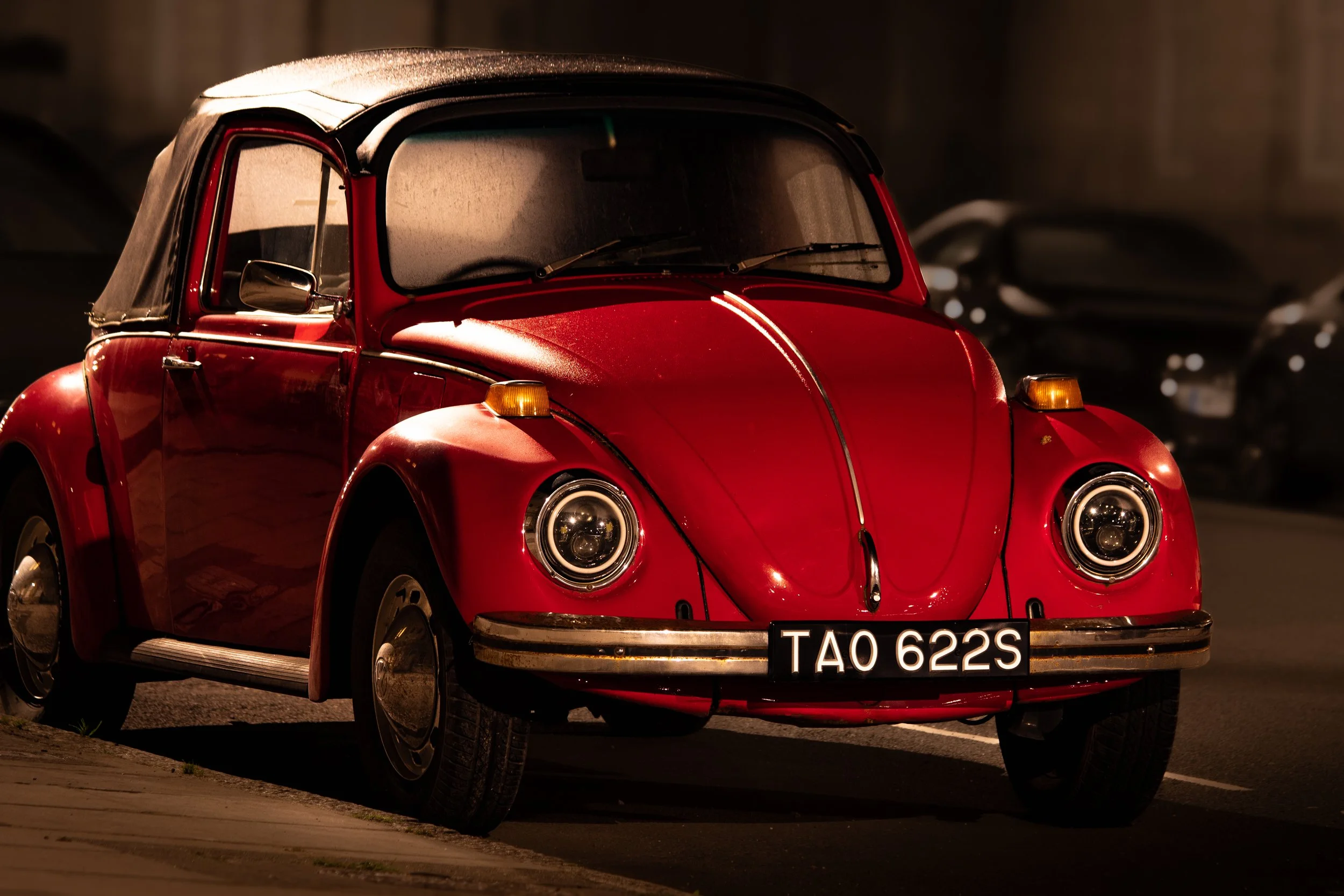 A red vintage Volkswagen Beetle with a black convertible top parked on a dark street at night, featuring a black license plate with white characters that reads 'TAO 622S'.
