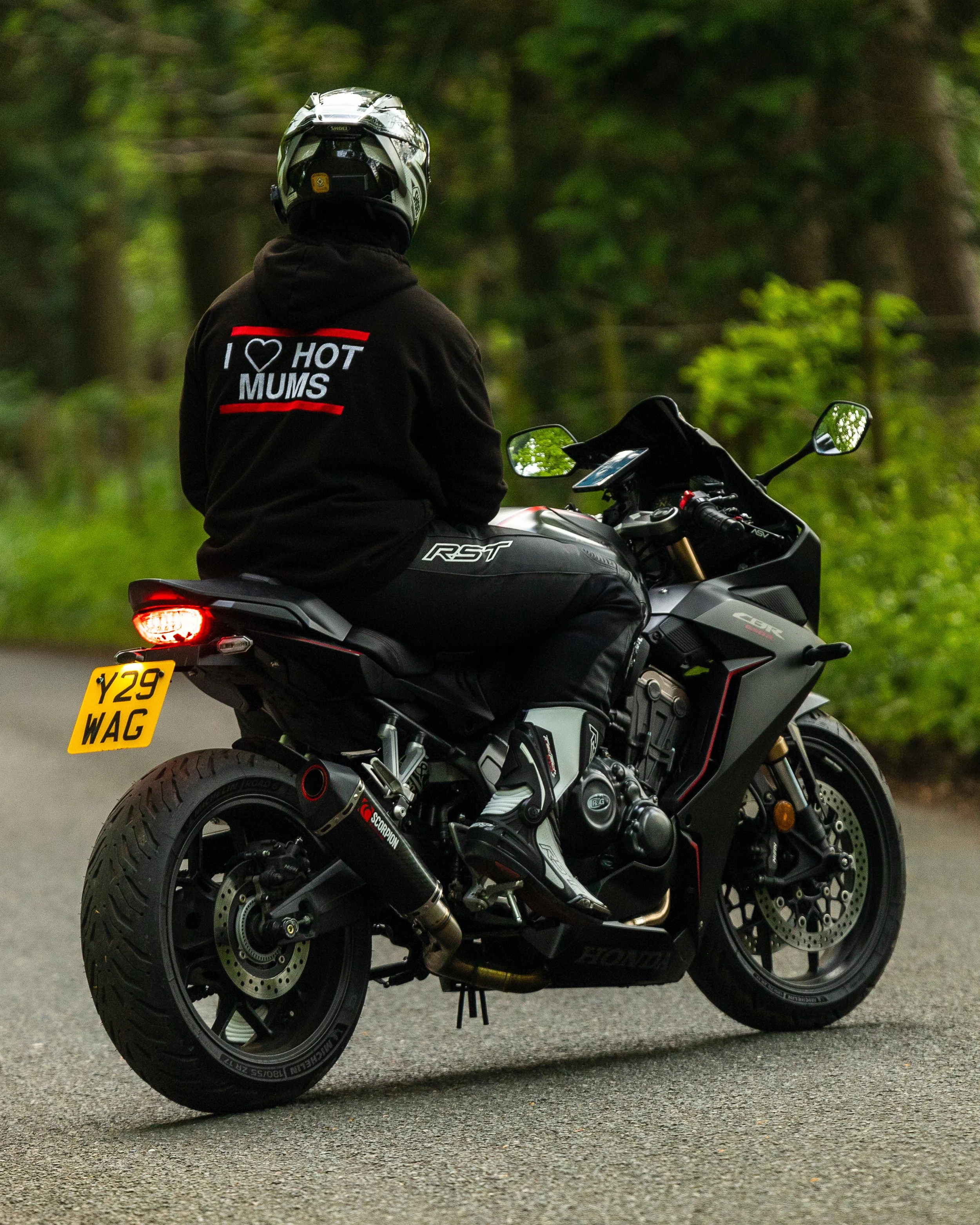 Person wearing a helmet and a hoodie with the words "I love hot mums" printed on the back, sitting on a black sports motorcycle on a paved road surrounded by green trees.