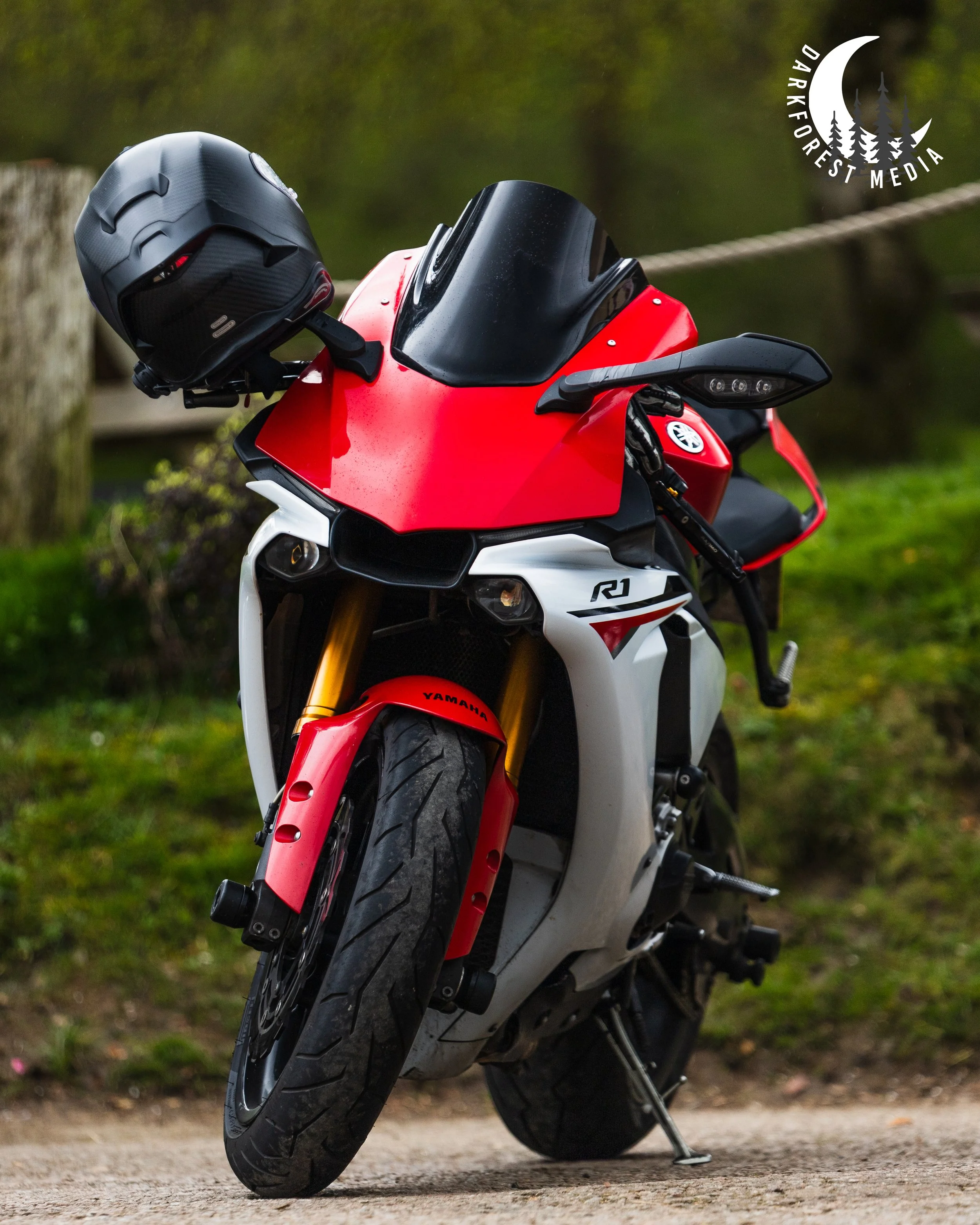 A red and white Yamaha R1 motorcycle parked on a dirt ground with a black helmet resting on the handlebar, surrounded by greenery and trees in the background.