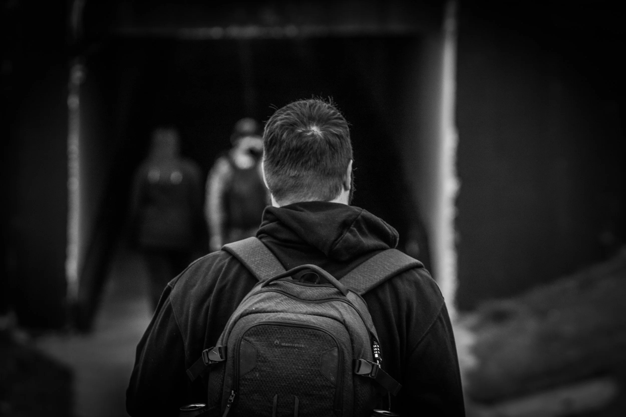 A person with a backpack facing away, with other people blurred in the background, in a black-and-white photo.