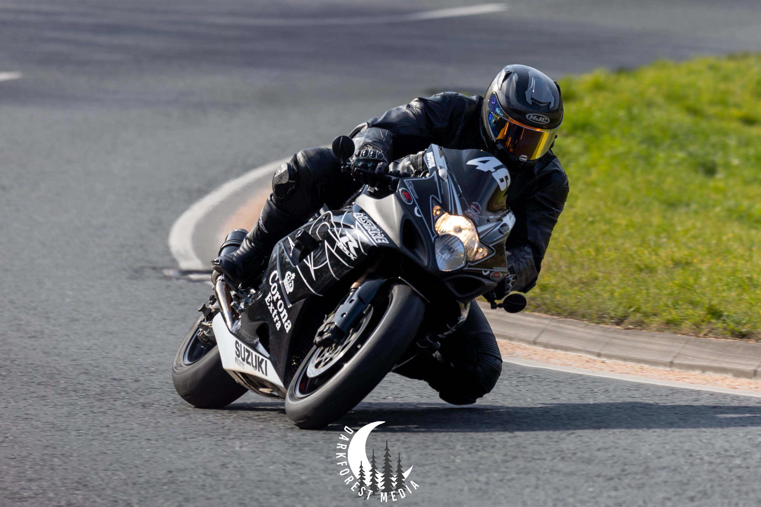 A motorcyclist wearing black protective gear and a helmet, leaning into a turn on a winding road with green grass on the side.
