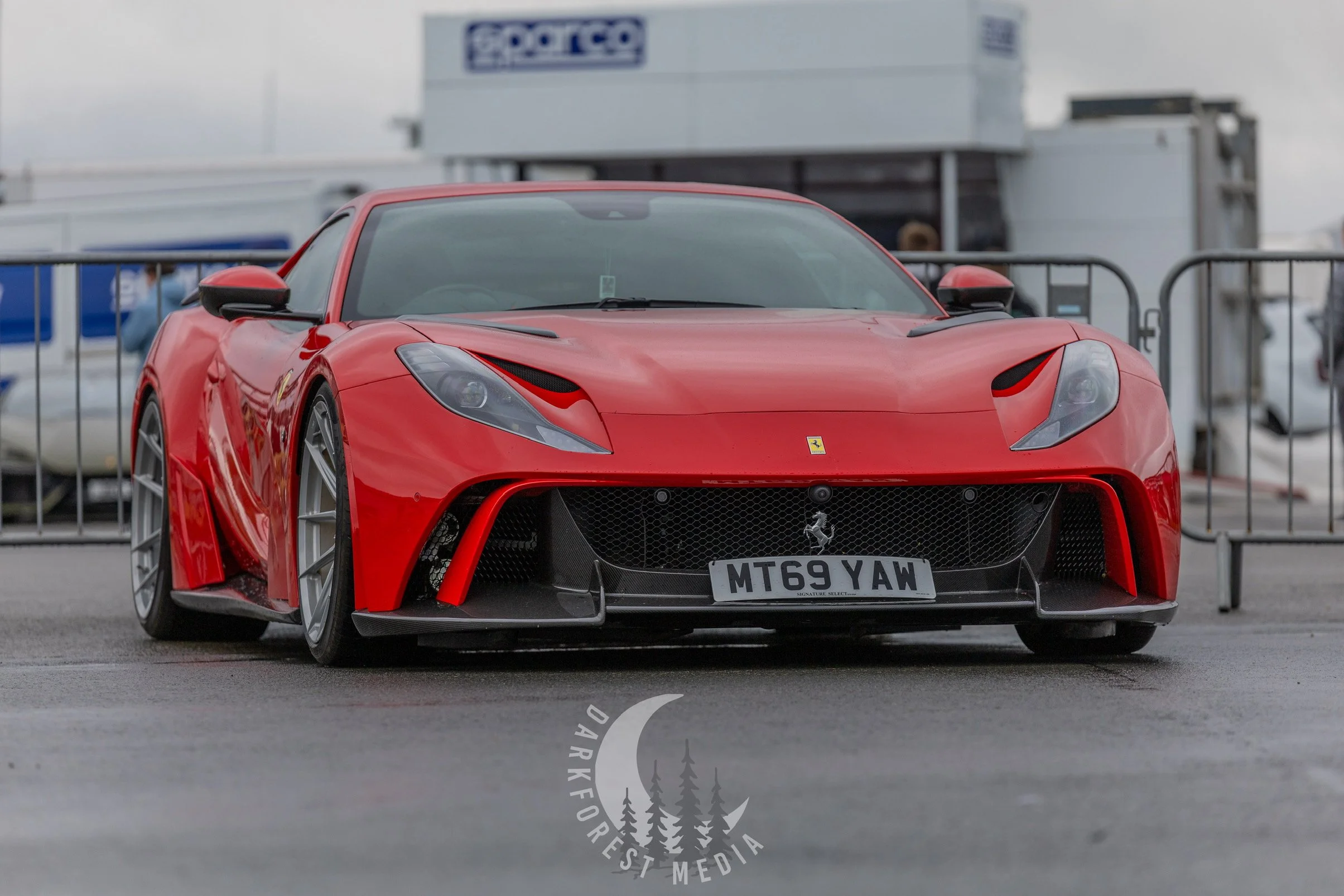 Red Ferrari sports car with black accents, parked outdoors at a car event. The car has a low, aerodynamic design, distinctive headlights, and a license plate reading 'MT69 YAW.' In the background, there are other vehicles and event barriers.