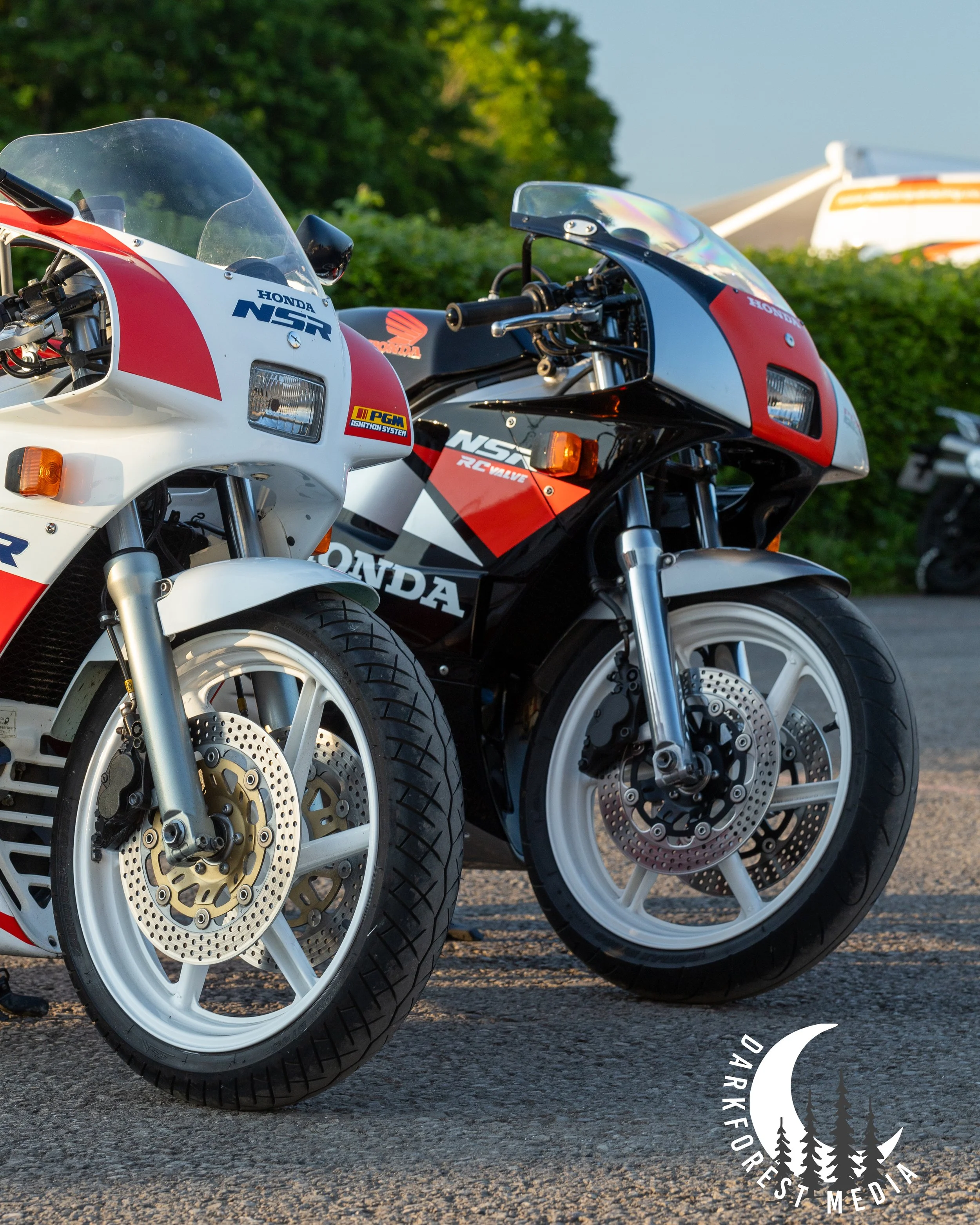 Close-up of two vintage Honda motorcycles parked on a paved surface with trees in the background, featuring detailed front wheels, disc brakes, and windshields.