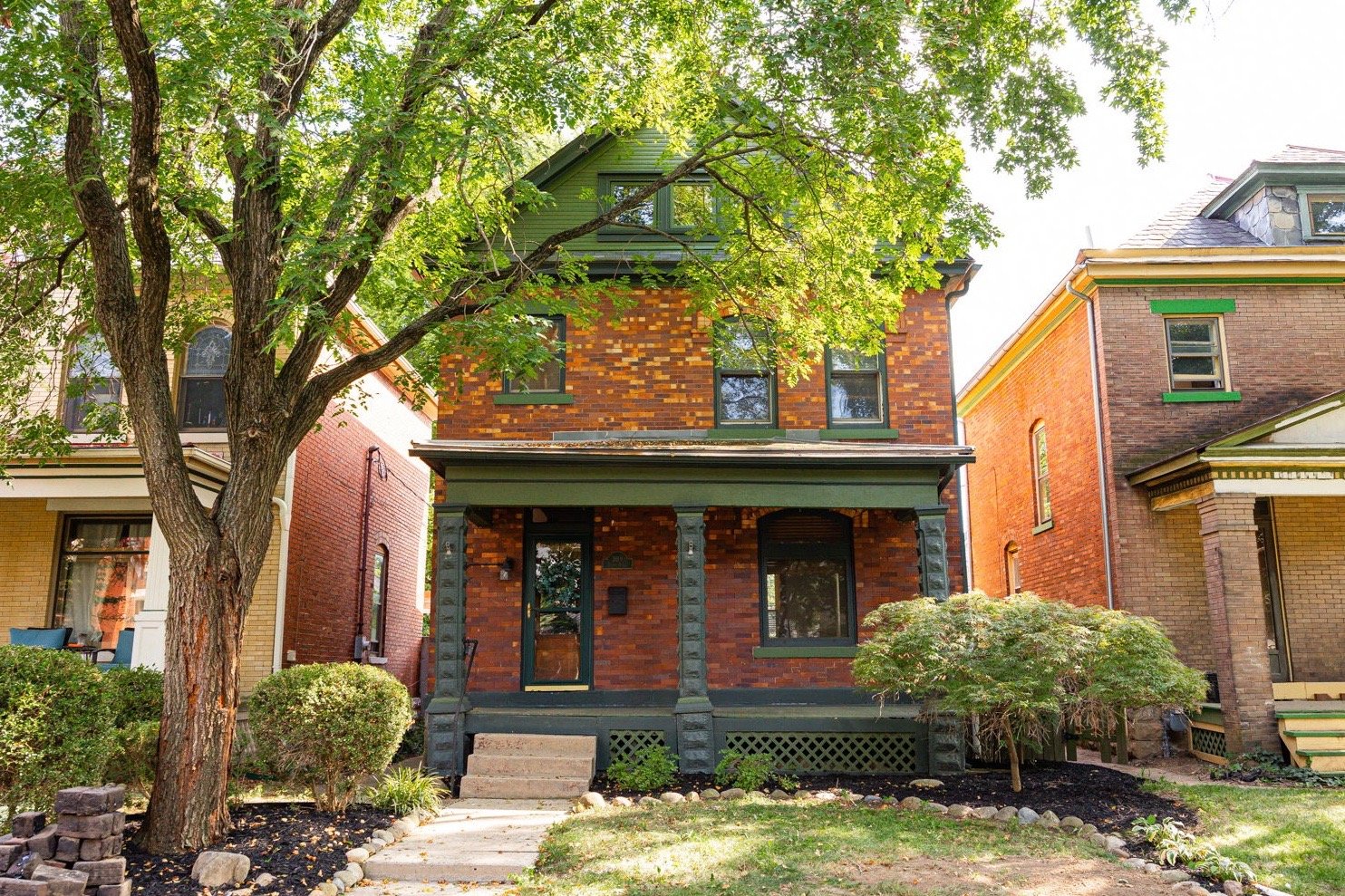 A three-story brick house with a green roof, green window frames, and a green front porch. The house is surrounded by trees and shrubs, with a sidewalk leading to the front steps.